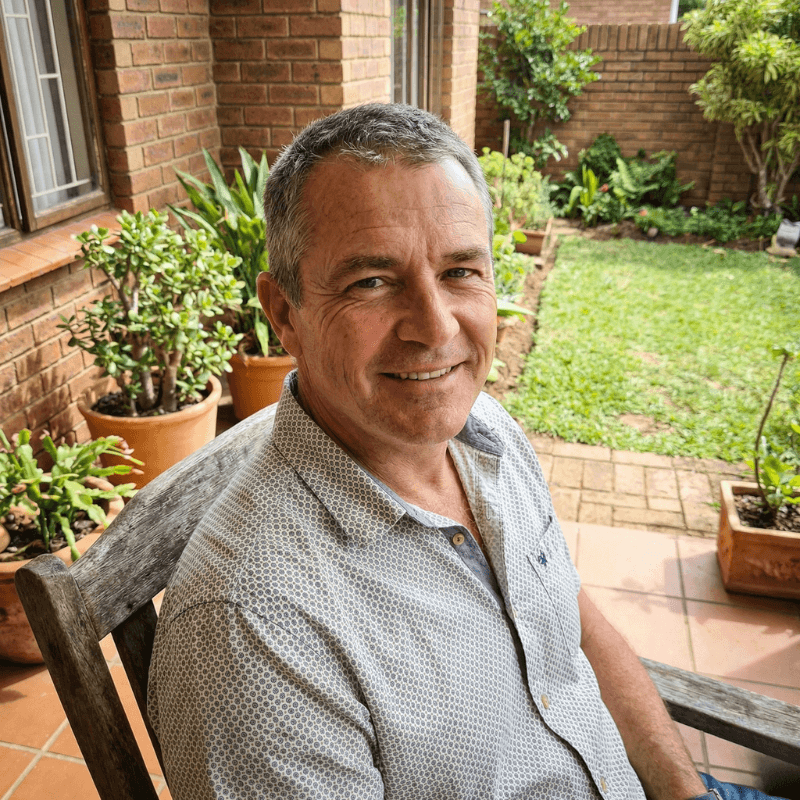 A middle-aged man with a beard, wearing a denim shirt and a leather vest, smiling outdoors in front of a white house with a landscaped yard.