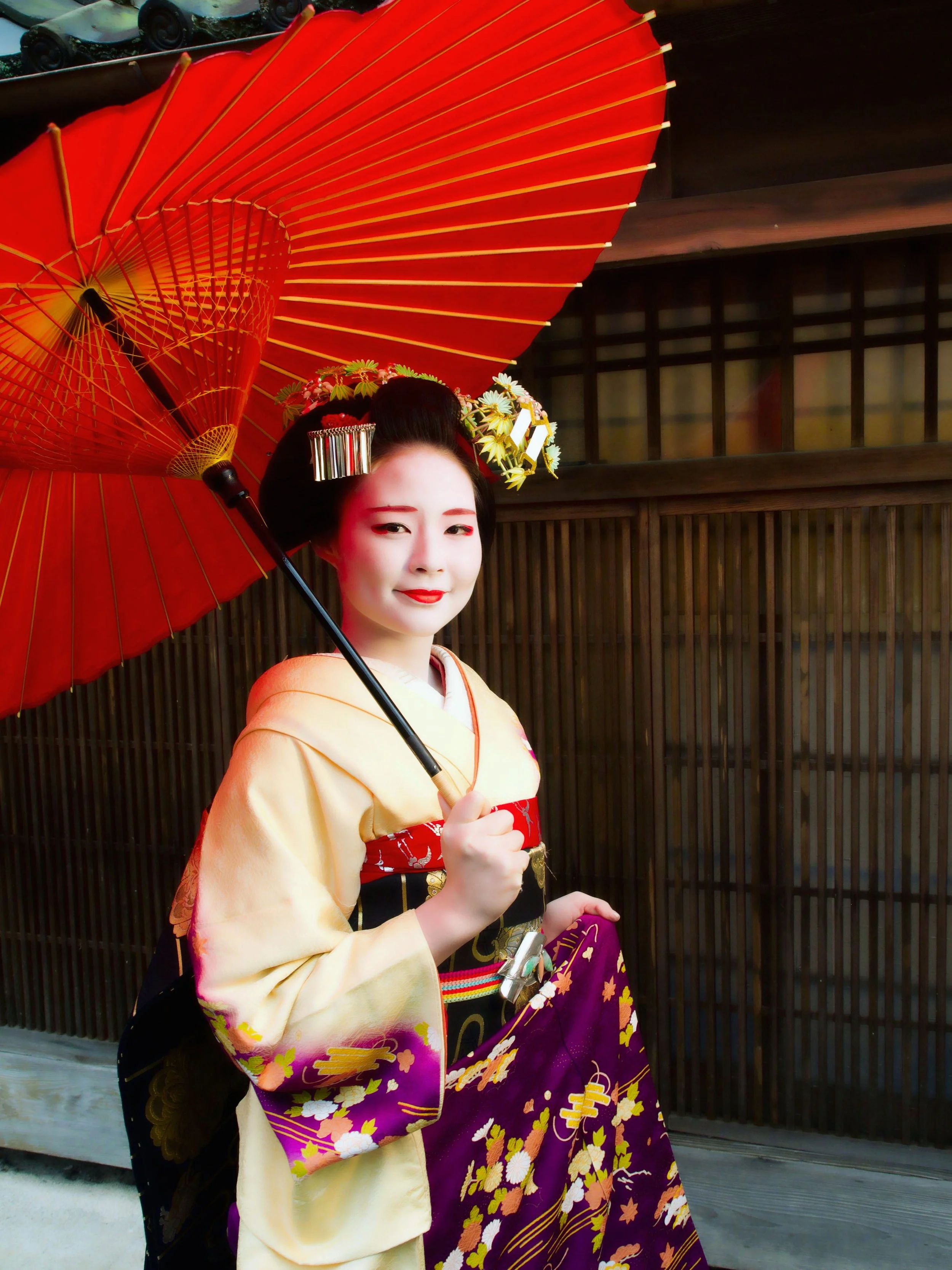 Une femme en kimono traditionnel japonais, tenant un parapluie rouge avec des motifs colorés, en face d'un mur en bois.