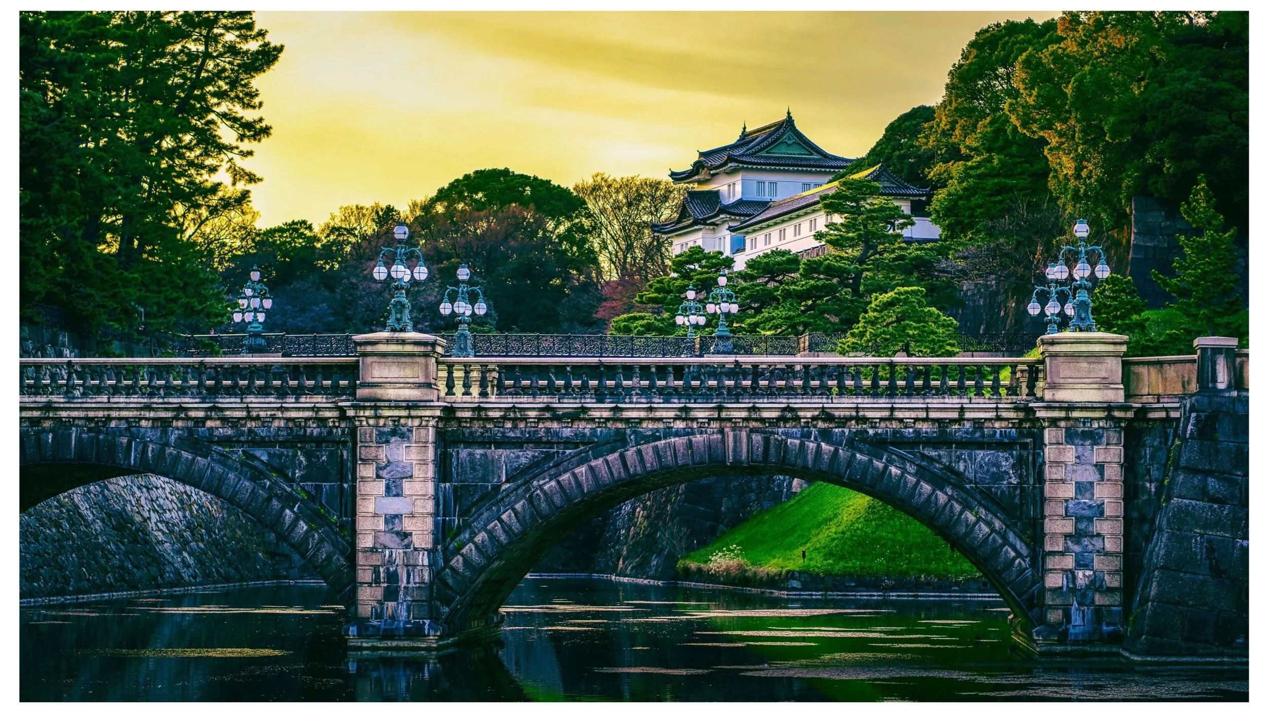 Un pont en pierre avec des lampadaires en fer forgé en premier plan, un bâtiment traditionnel japonais avec un toit en tuiles noires et une structure blanche en arrière-plan, entouré d'arbres verts et un ciel jaune au coucher du soleil.