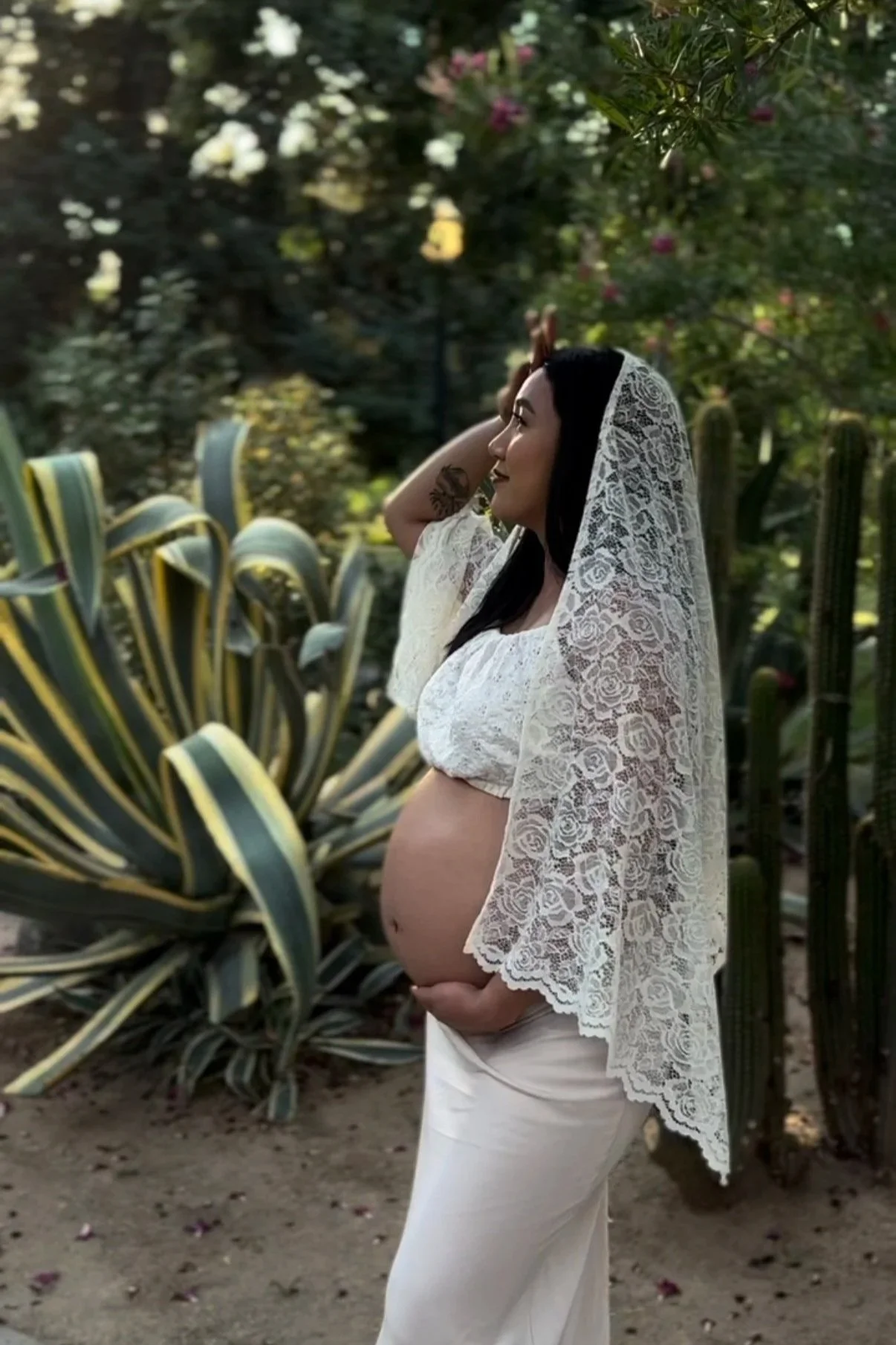 A pregnant woman stands outdoors in a garden, wearing a white lace shawl and a white top, with greenery and cacti in the background.