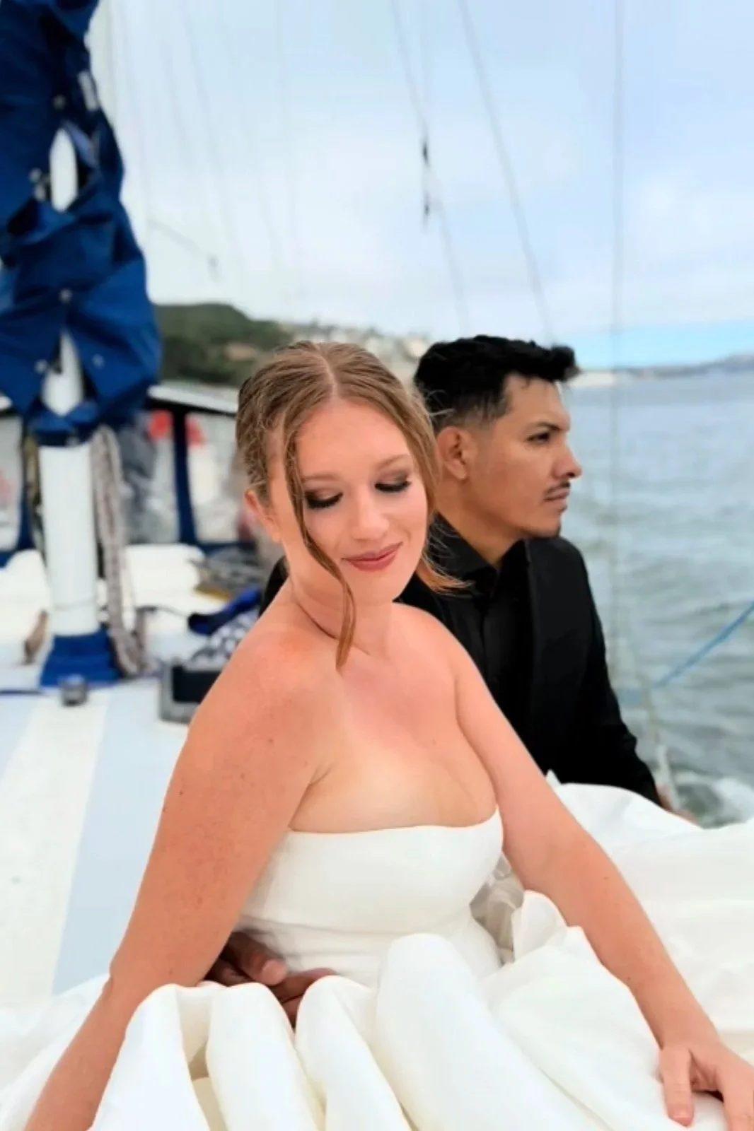 A woman in a white strapless dress sitting on a boat with a man in a black shirt behind her, near the water with a blurred harbor and sky in the background.