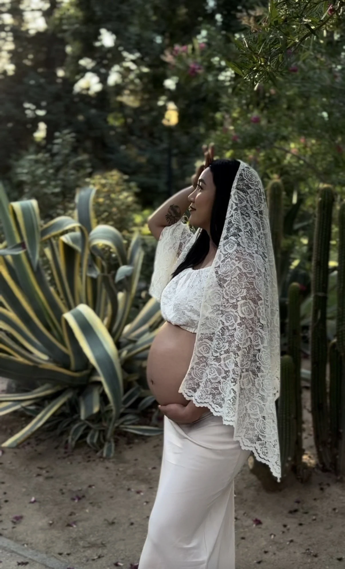 A pregnant woman stands outdoors in a garden, wearing a white lace shawl and a white top, with greenery and cacti in the background.