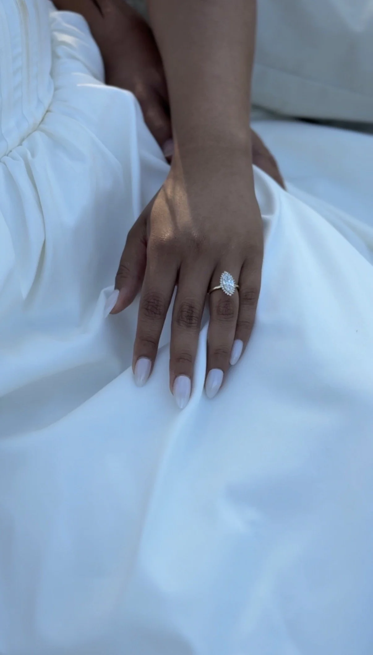 Close-up of a woman's hand resting on white fabric, showcasing a large oval diamond engagement ring with smaller diamonds around it.