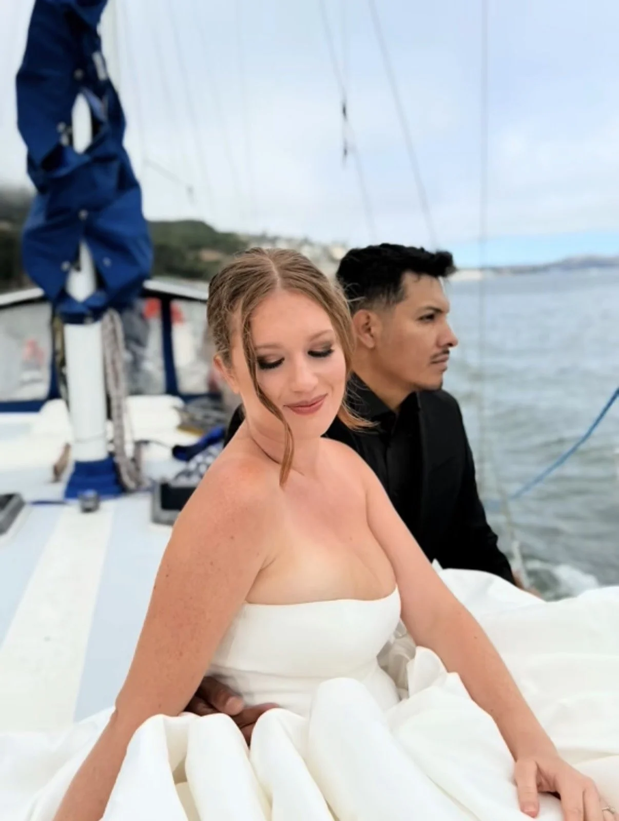 A woman in a white strapless dress sitting on a boat with a man in a black shirt behind her, near the water with a blurred harbor and sky in the background.