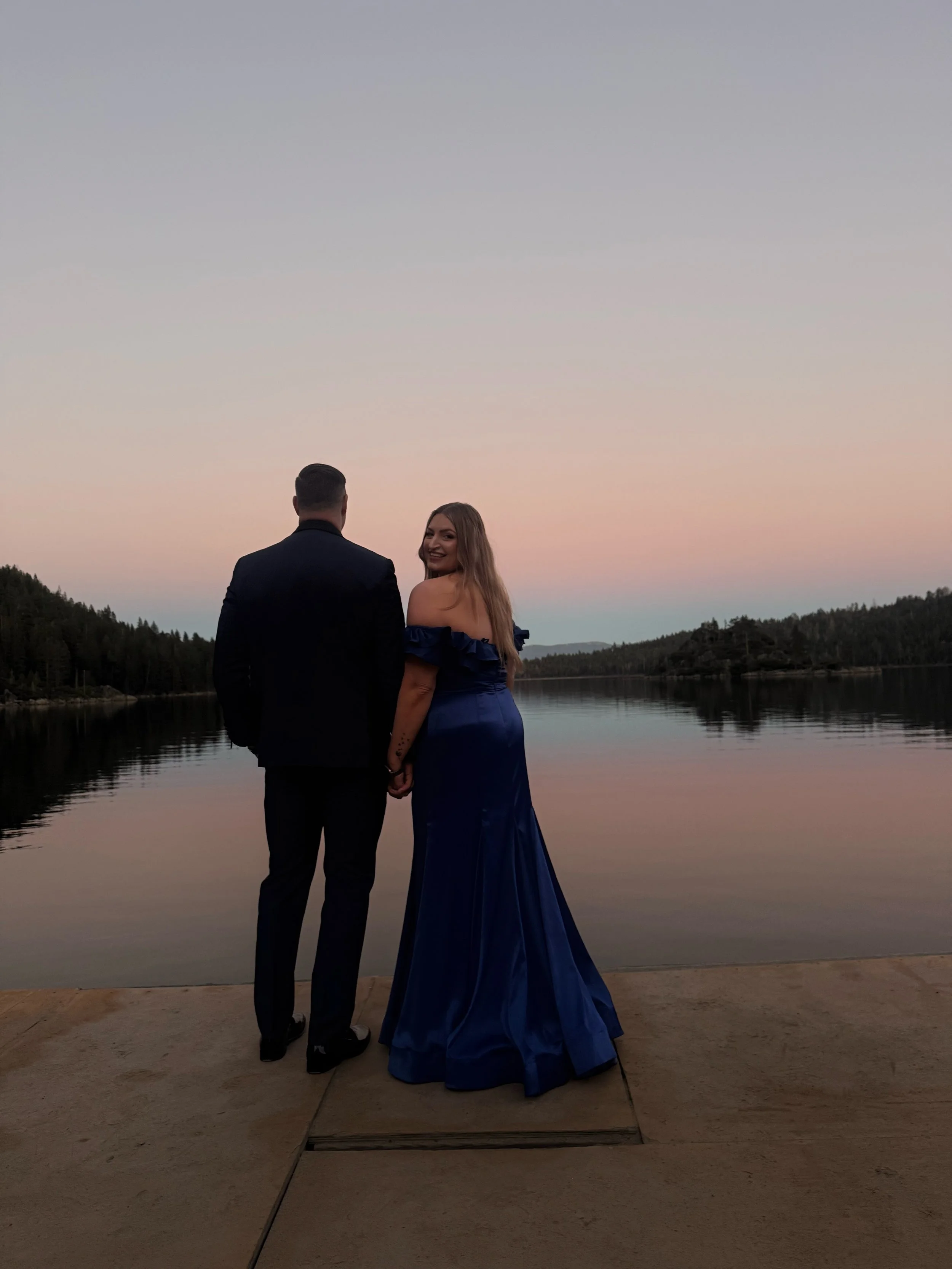 A couple holding hands, standing on a dock by a calm lake at sunset. The woman is wearing a long, blue gown, and the man is dressed in a black suit. She is smiling and looking over her shoulder at the camera.