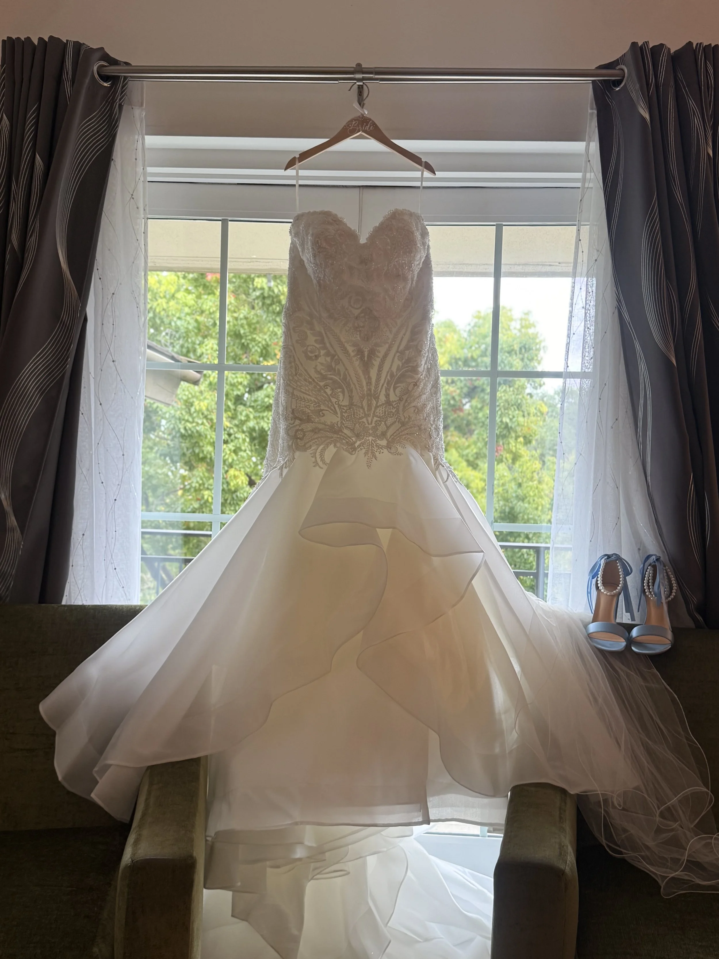 Elegant white wedding gown with lace bodice and full tulle skirt, hanging on a wooden hanger near a window, with a pair of blue high-heeled shoes nearby.