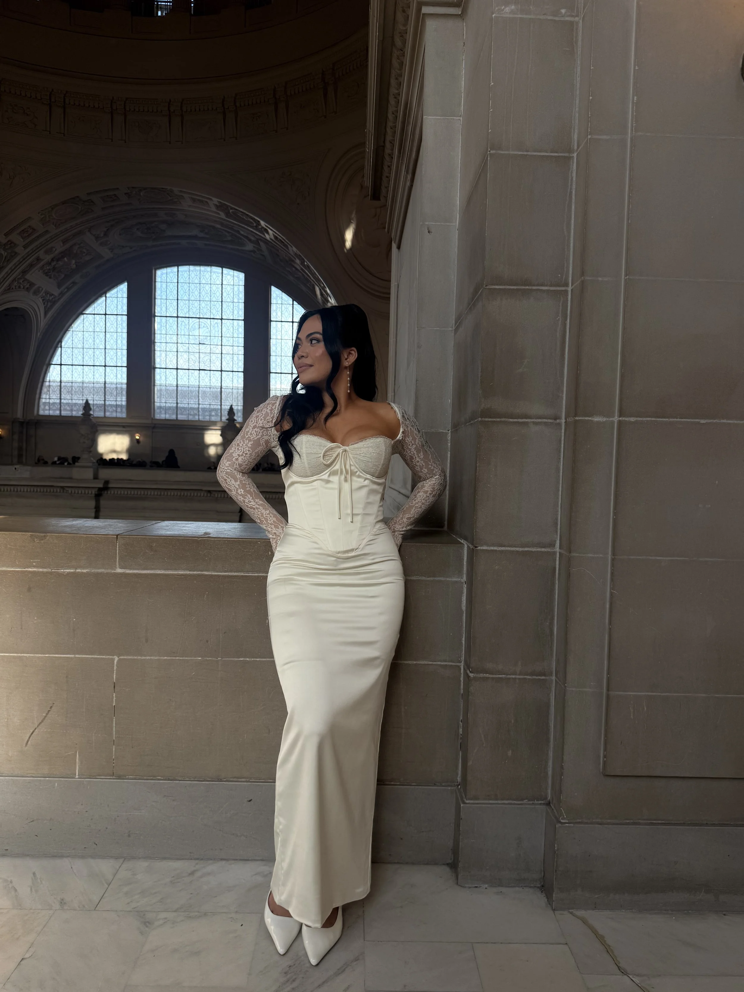 A woman in an elegant cream-colored dress with lace sleeves and pointed heels, standing inside a grand, historic building with large arched windows and ornate architectural details.