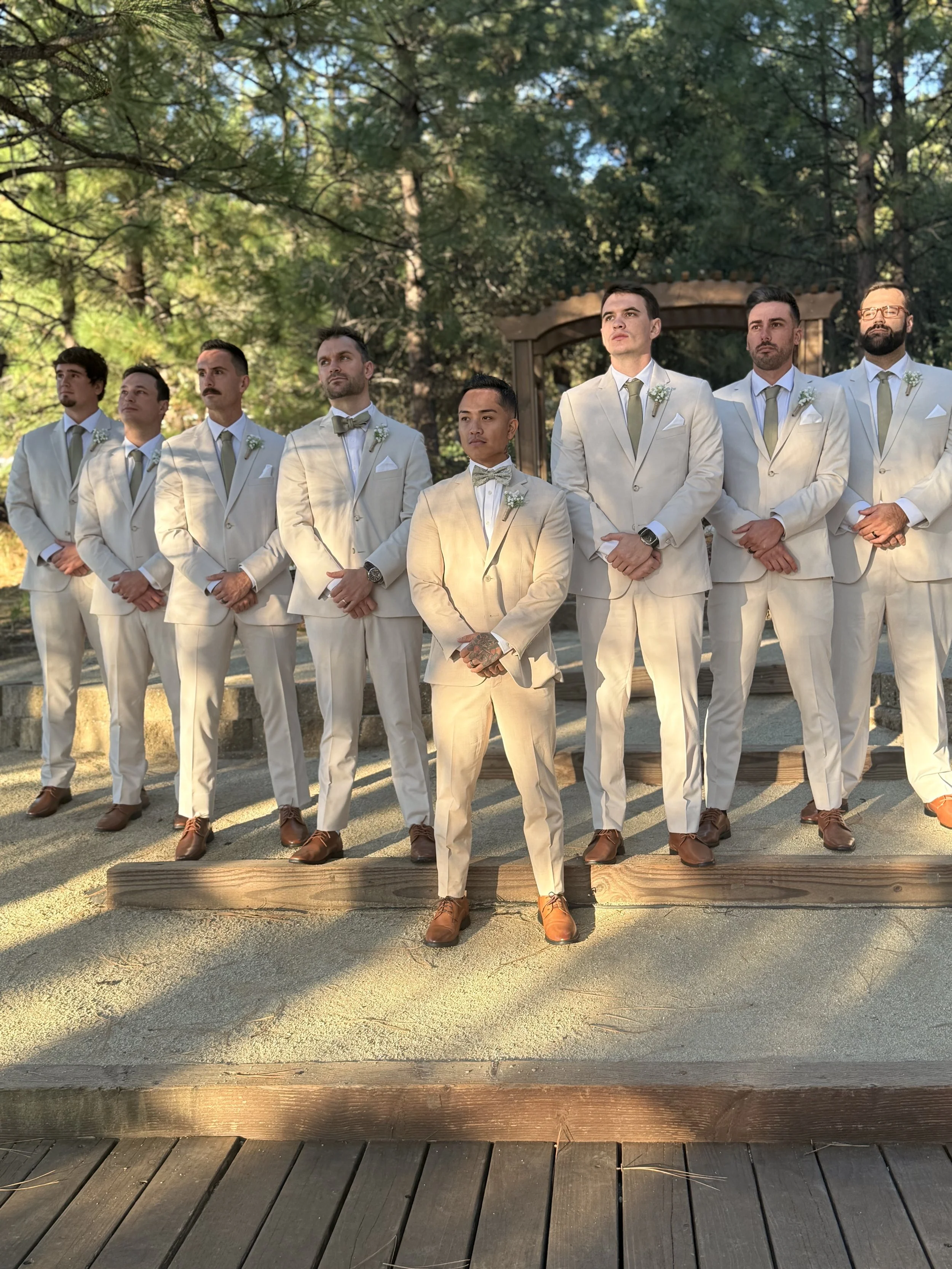 A groom and seven groomsmen standing outdoors during a wedding, all dressed in matching white suits with light green ties, in front of a natural wooded background with trees and sunlight.