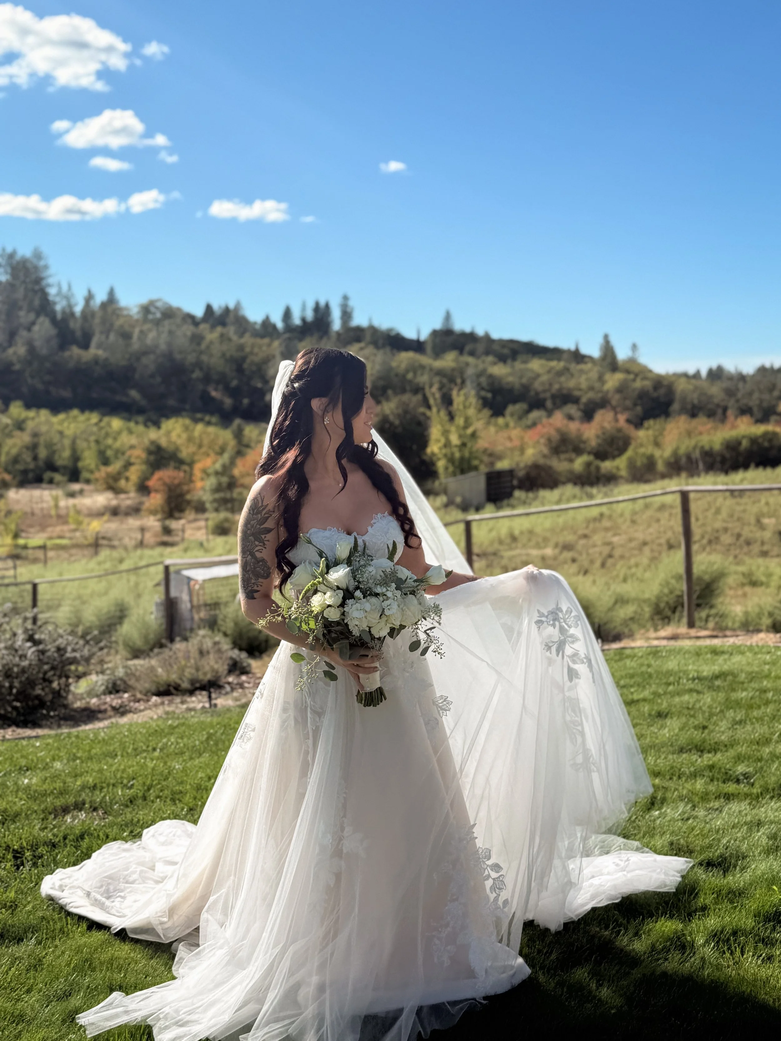 Bride in a white wedding gown holding a bouquet of white flowers, standing outdoors on a grassy area under a blue sky with some clouds.