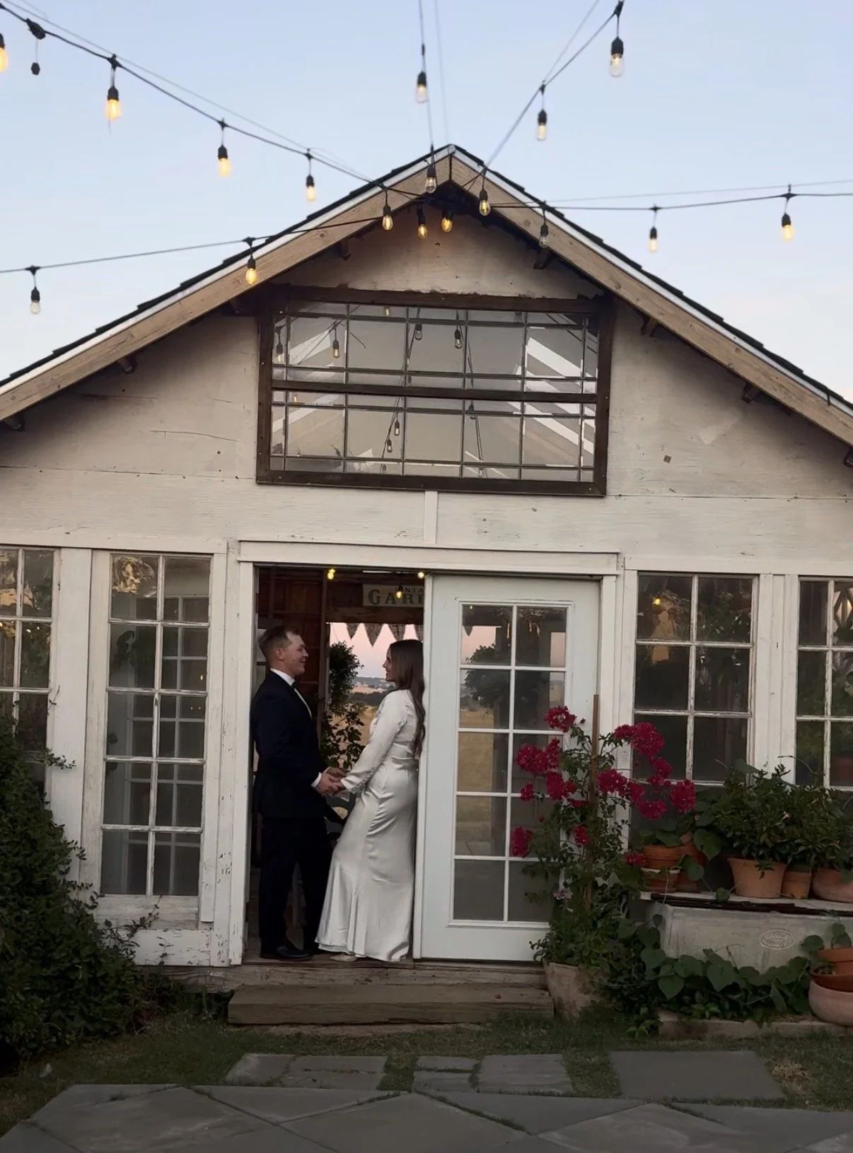 A couple in wedding attire holding hands and smiling at each other in front of a rustic greenhouse with string lights hanging above, during sunset.