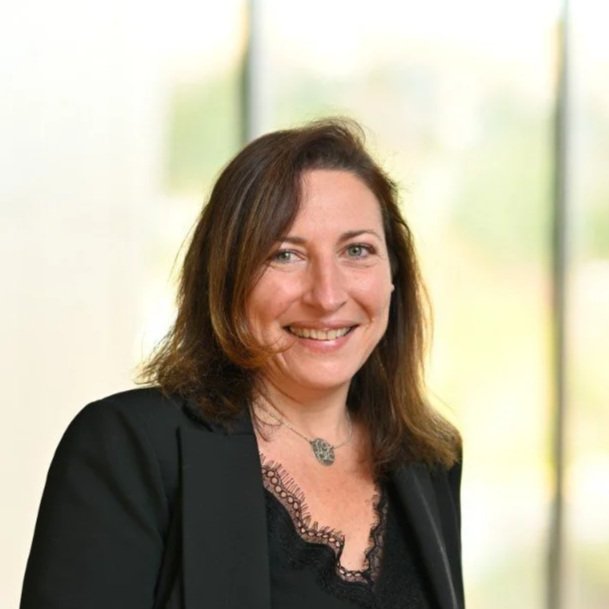 Portrait of a woman with brown hair smiling, wearing a black blazer and necklace, against a blurred background with windows.