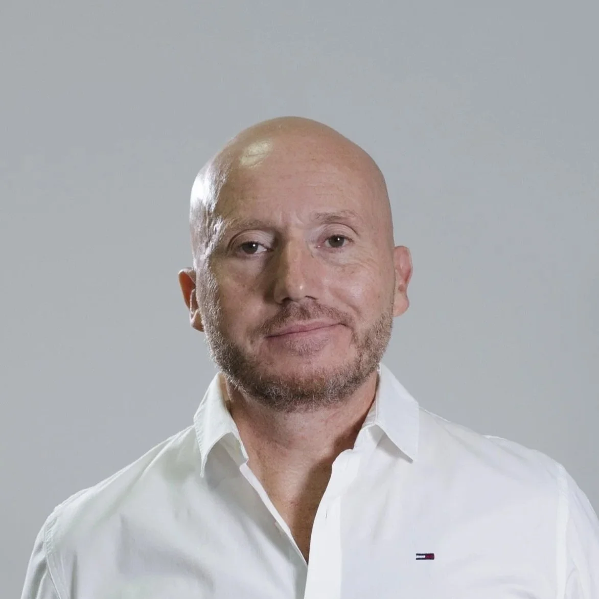 A middle-aged man with a bald head and beard, wearing a white shirt, looking at the camera against a plain gray background.