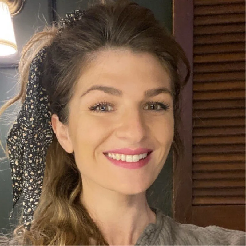 Close-up of a young woman with light skin and long wavy hair, smiling with visible teeth, in an indoor setting.