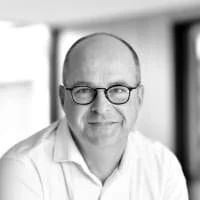 A black-and-white portrait of a middle-aged man with glasses, bald head, wearing a collared shirt, indoors.