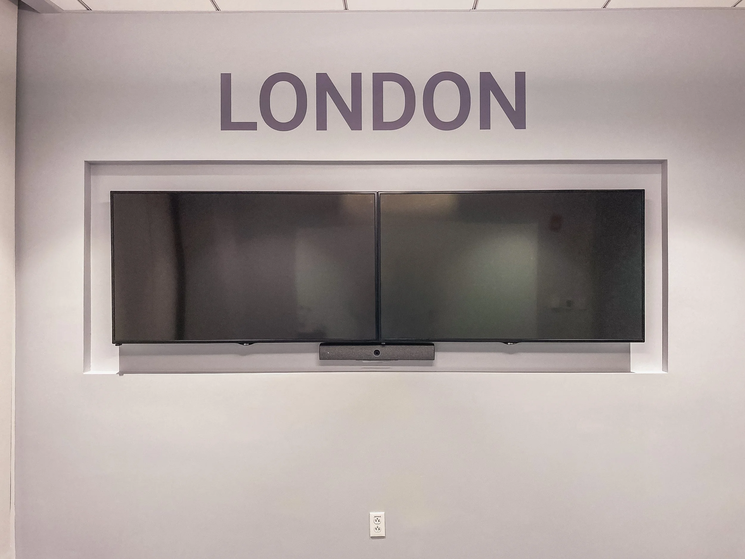 Large flat-screen TV mounted on a light gray wall with the word 'LONDON' written in large purple letters above. Below the TV, there is a power outlet with two sockets.