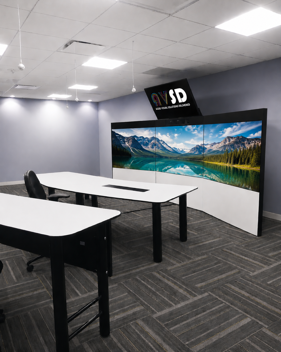 Empty office conference room with white tables, black chairs, a large curved multi-screen display showing a mountain lake landscape, and a smaller screen above displaying the logo 'RV SD Audio Visual Solutions Delivered.'