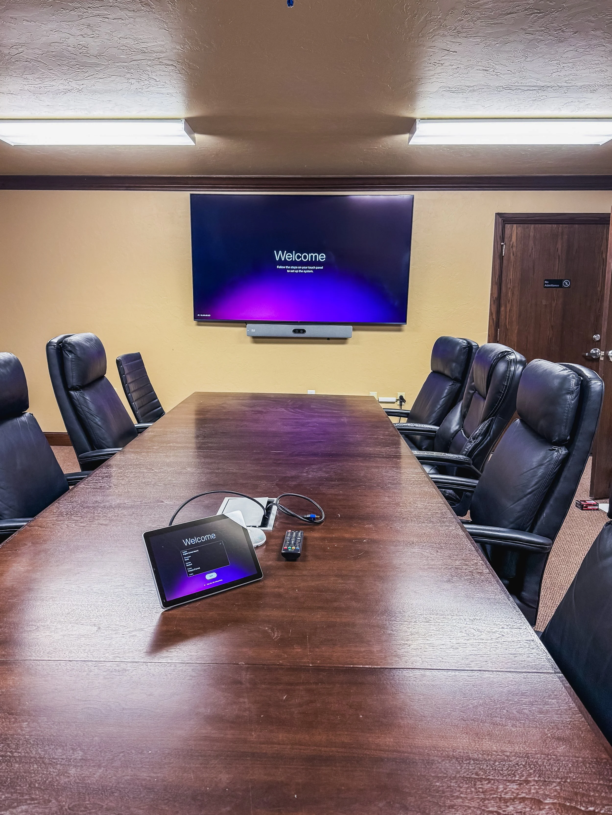 A conference room with a large wooden table, black leather chairs, a wall-mounted screen displaying a welcome message, a tablet and remote control on the table, and a wooden door with no admittance sign.
