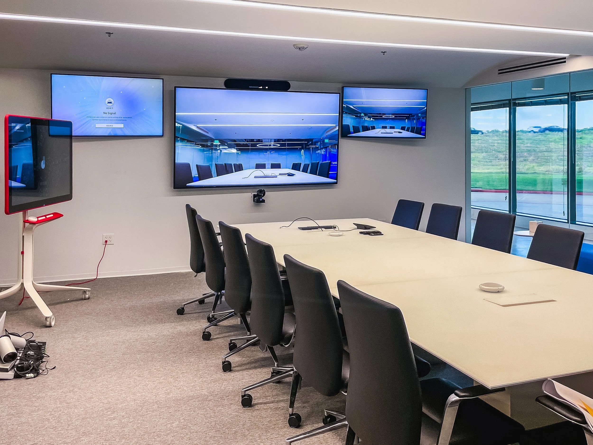 A conference room with a long white table, black chairs, multiple wall-mounted screens, and large windows showing a grassy landscape outside.