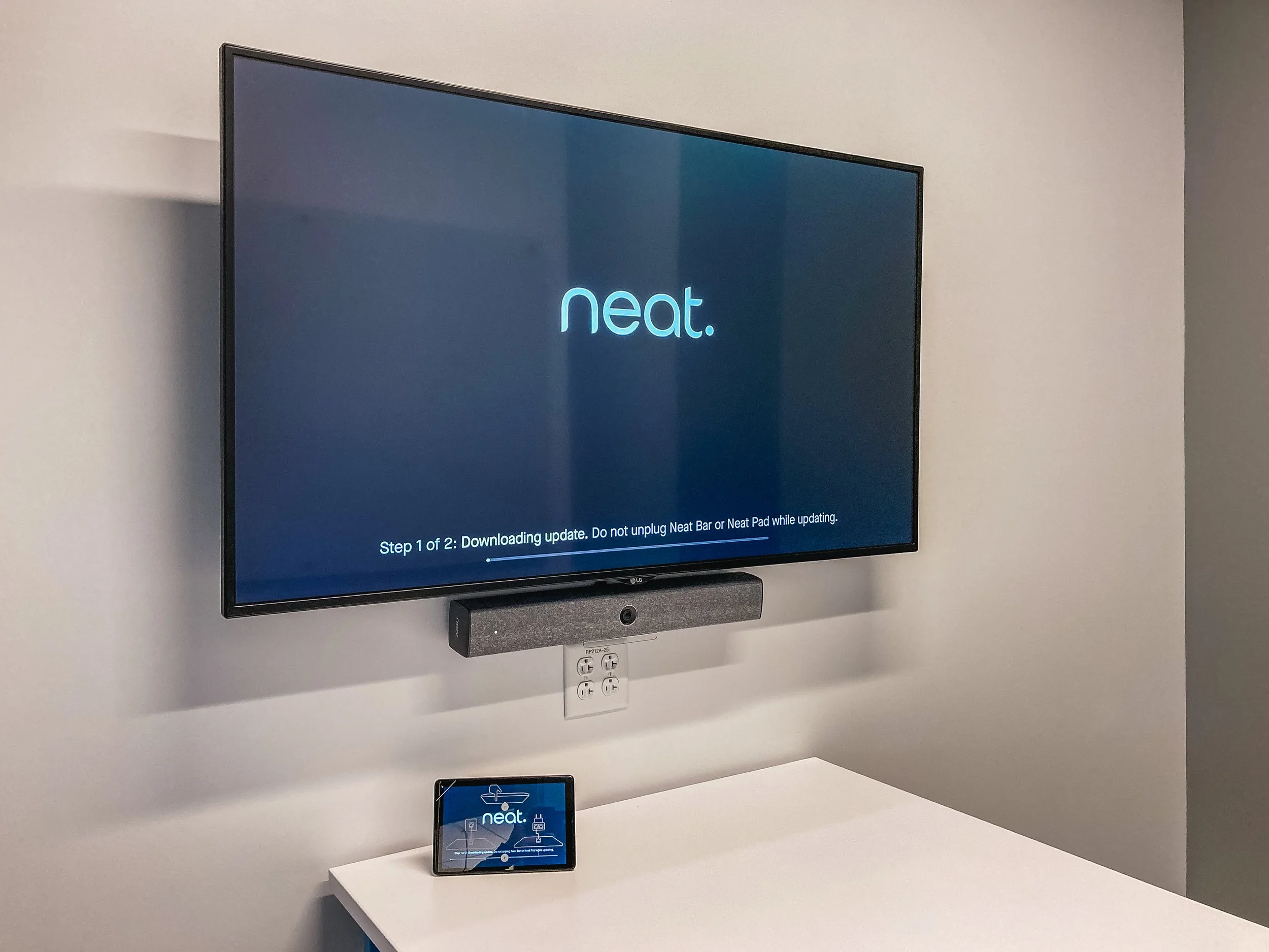 A television screen displays a software update message for Neat, a company that produces scanning and OCR devices. Below the screen, there is a small device with the Neat logo on it, placed on a white table, and a door with multiple electrical outlet