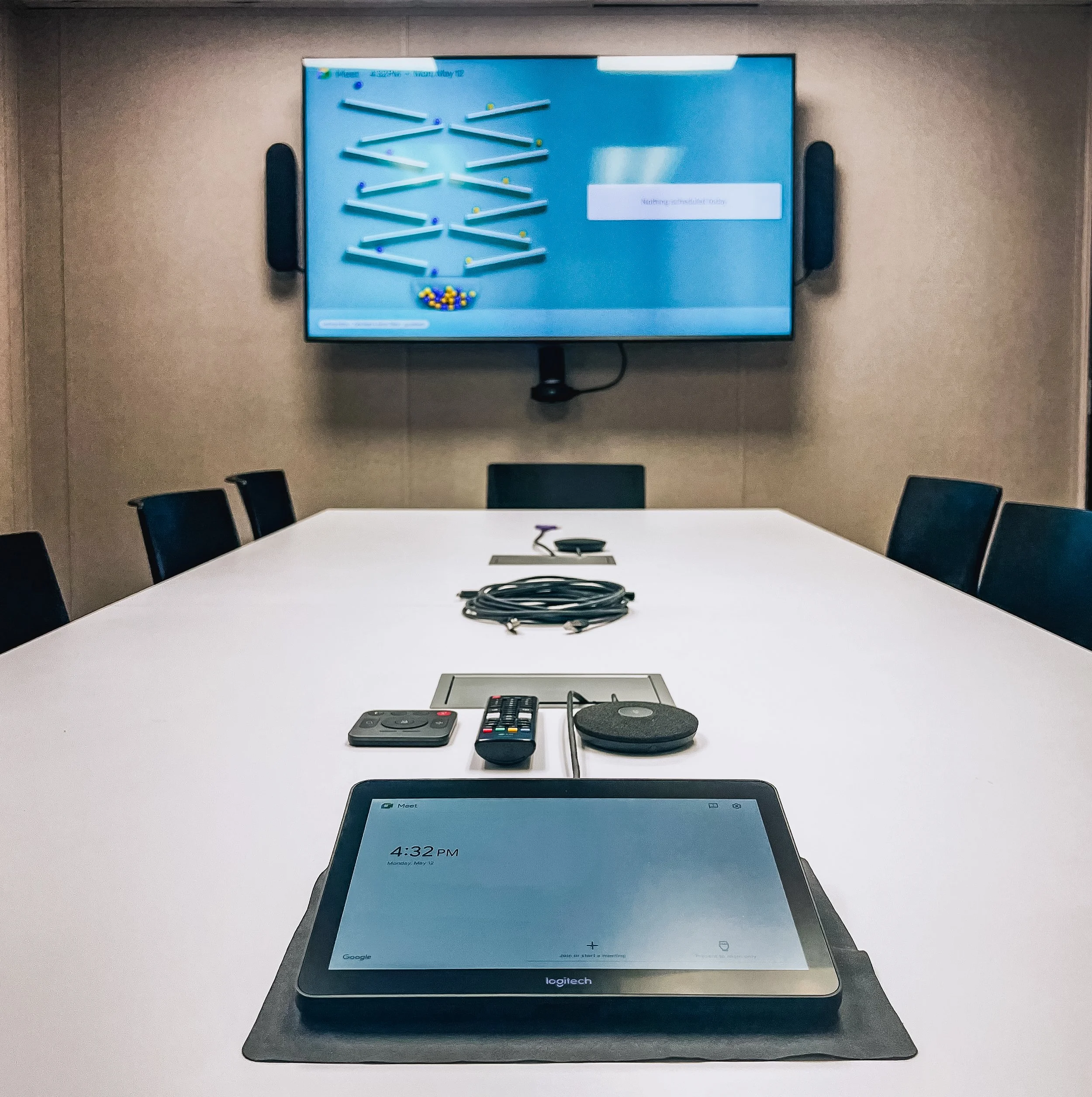 A conference room table with electronic devices including a black remote control, a black pad, and a black-and-silver card reader. The background shows a wall-mounted digital display and chairs around the table.