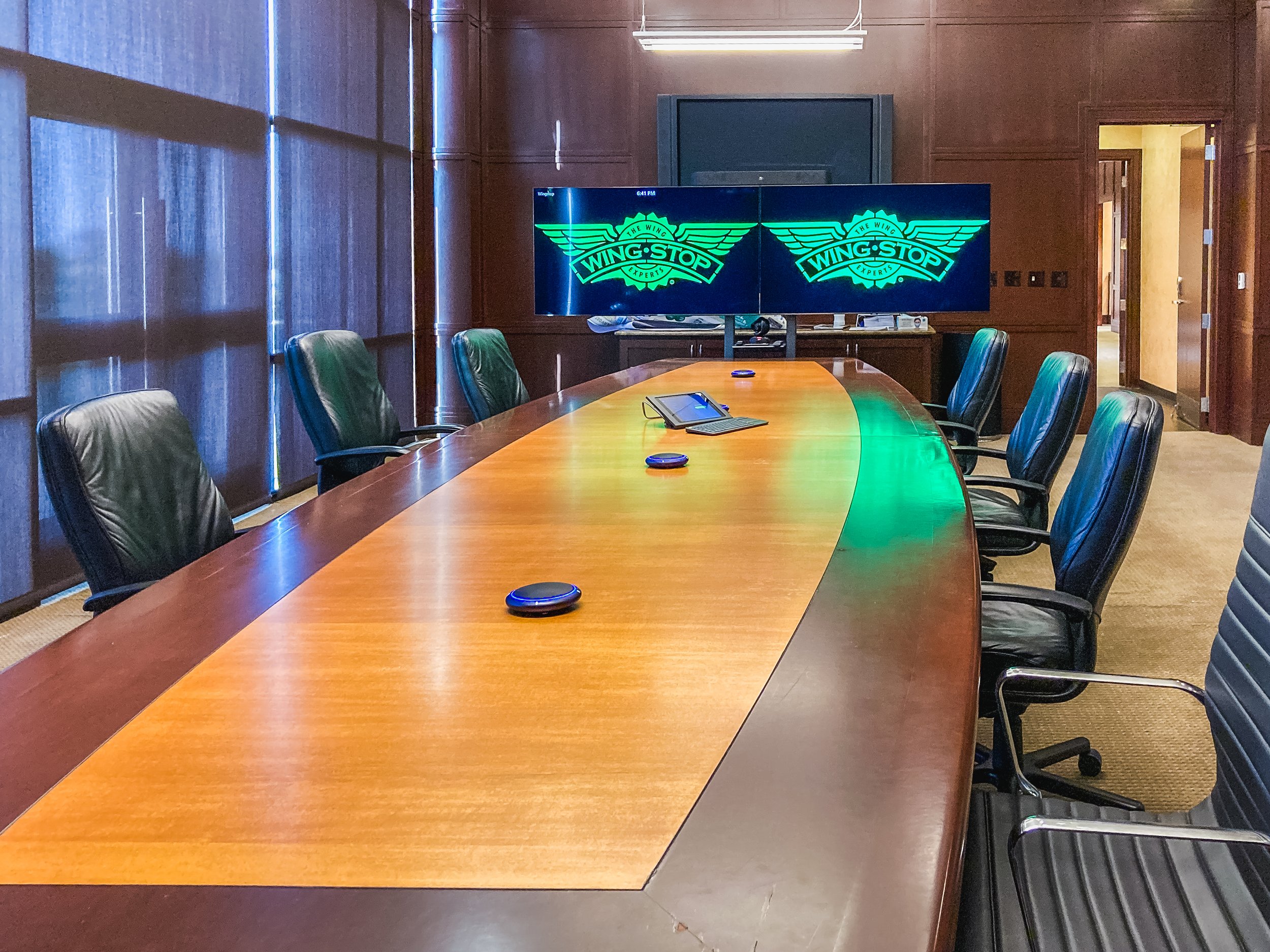A large conference room with a long wooden table, black leather chairs, two computer mice, and a conference phone. There is a dual-screen monitor with the Wing Stop logo on it, and a wood-paneled wall with a doorway leading to a brightly lit hallway.