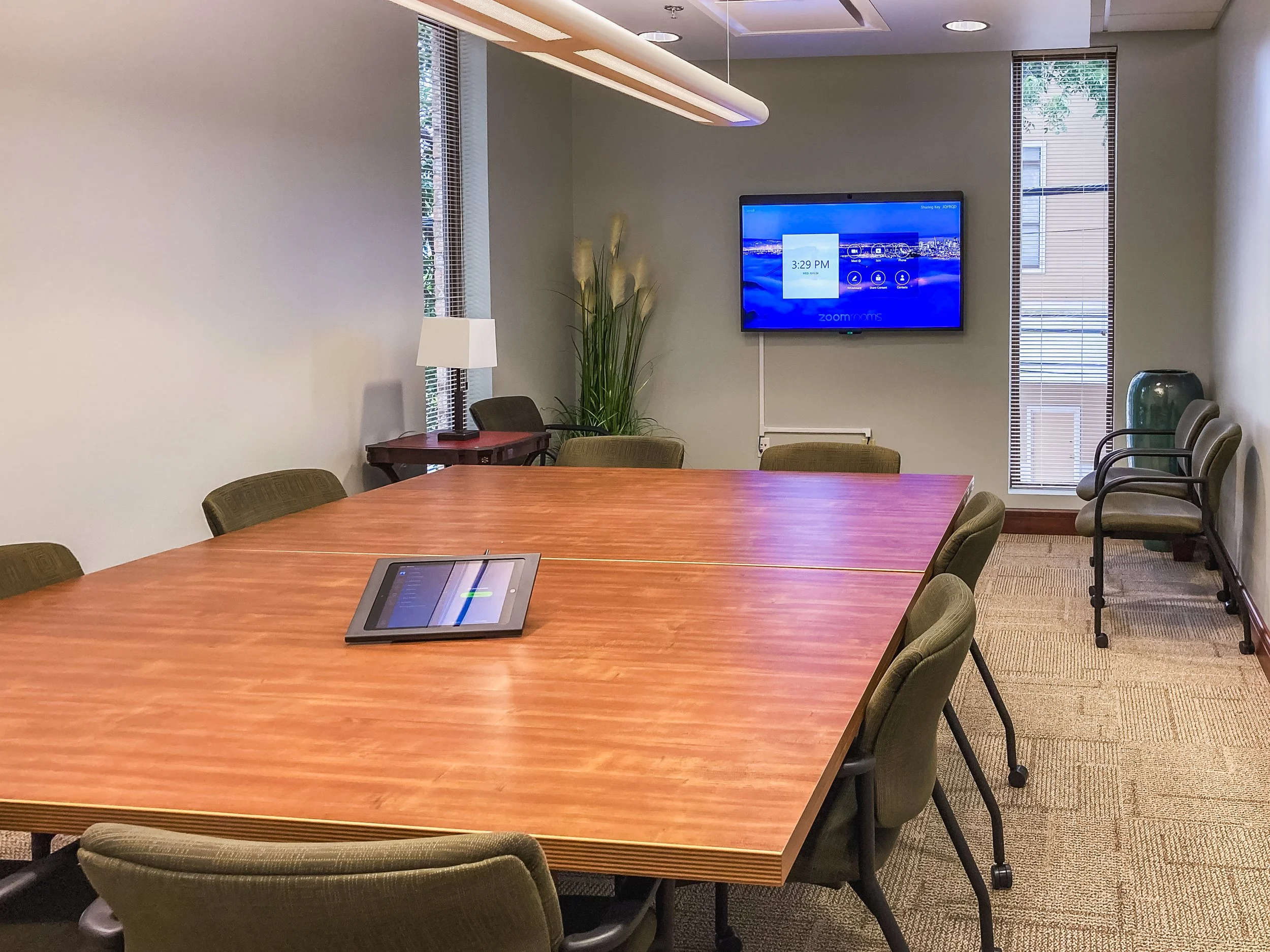 A conference room with a large wooden table surrounded by green upholstered chairs. A tablet device is on the table. There is a wall-mounted screen showing the time 3:29 PM and some digital interface options. The room has windows with blinds, a side 