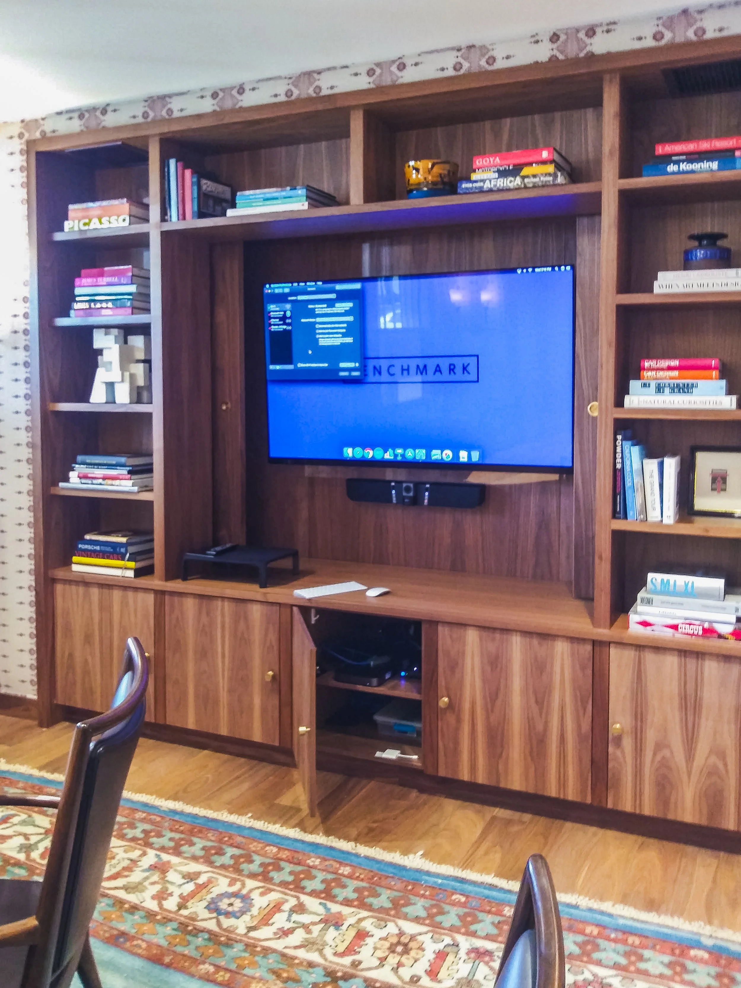Wooden wall unit with shelves filled with books, a television, and decorative objects in a living room with a patterned rug and chairs.
