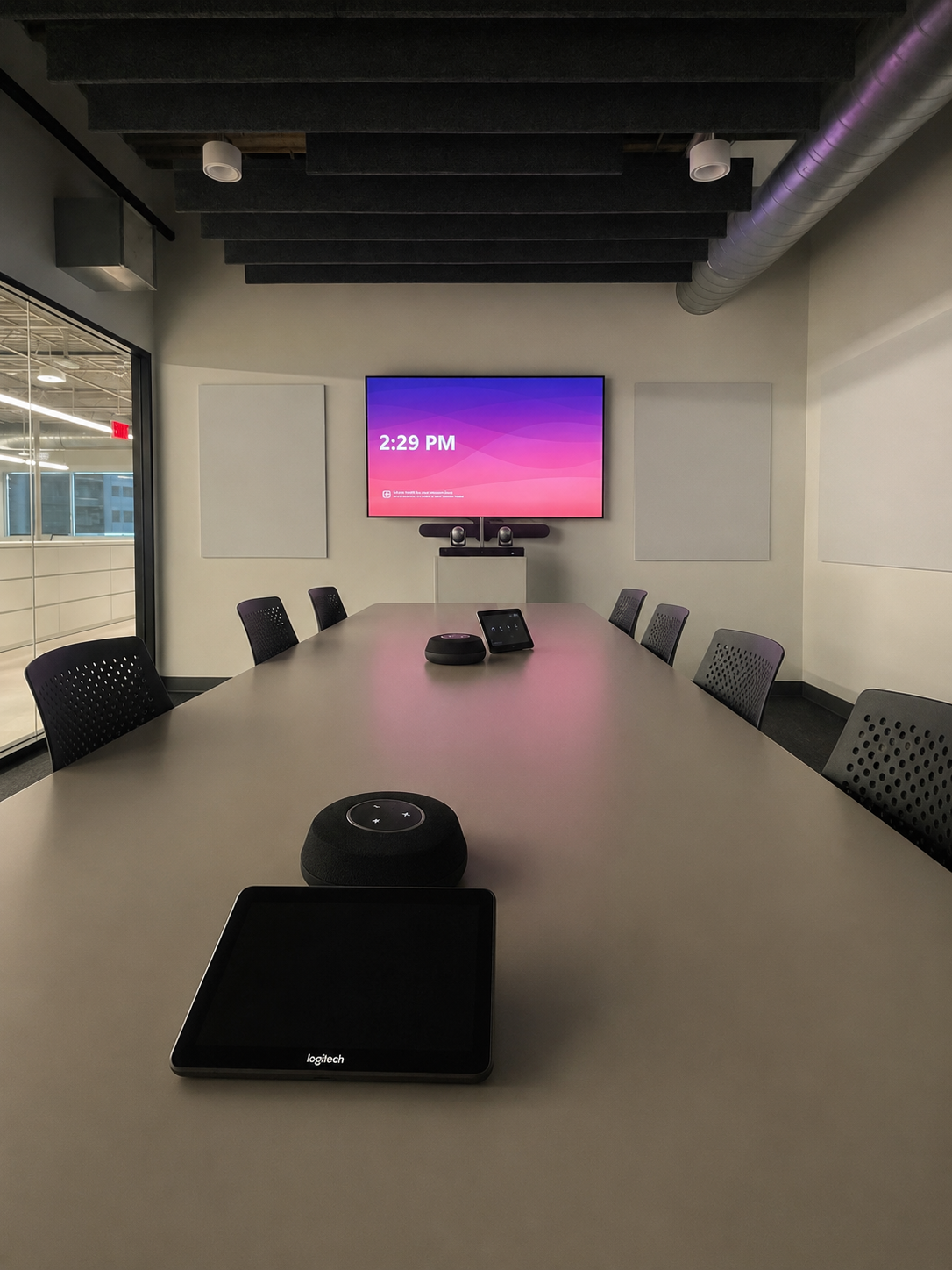 Conference room with a large table, black chairs, a wall-mounted screen displaying the time 2:29 PM, video conferencing equipment, and a tablet on the table. There is a glass wall to the left showing an adjacent room and ceiling ductwork overhead.