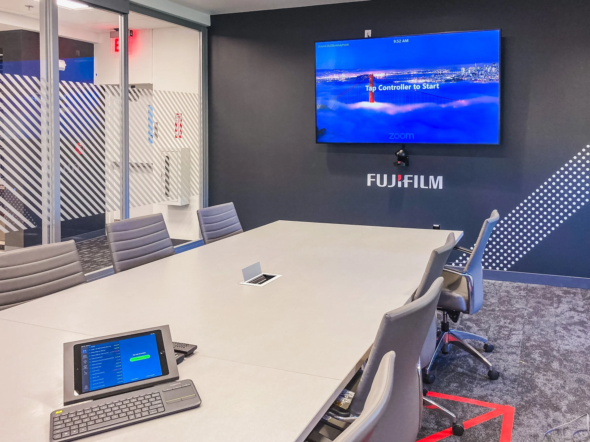 A conference room with a long table surrounded by gray chairs. On the table, there is a laptop and a digital control panel. A large screen on the wall displays a blue background with an image of the Golden Gate Bridge and the words 'Tap Controller to