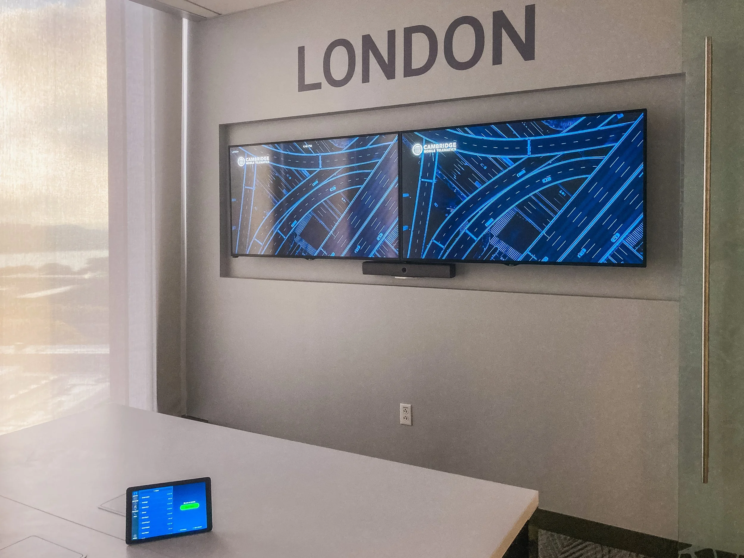 Two monitors displaying a blue digital map or circuit design with the text "Cambridge Mobile Telamatics" on the right monitor. The location tag "London" is visible on the top left of the monitors.