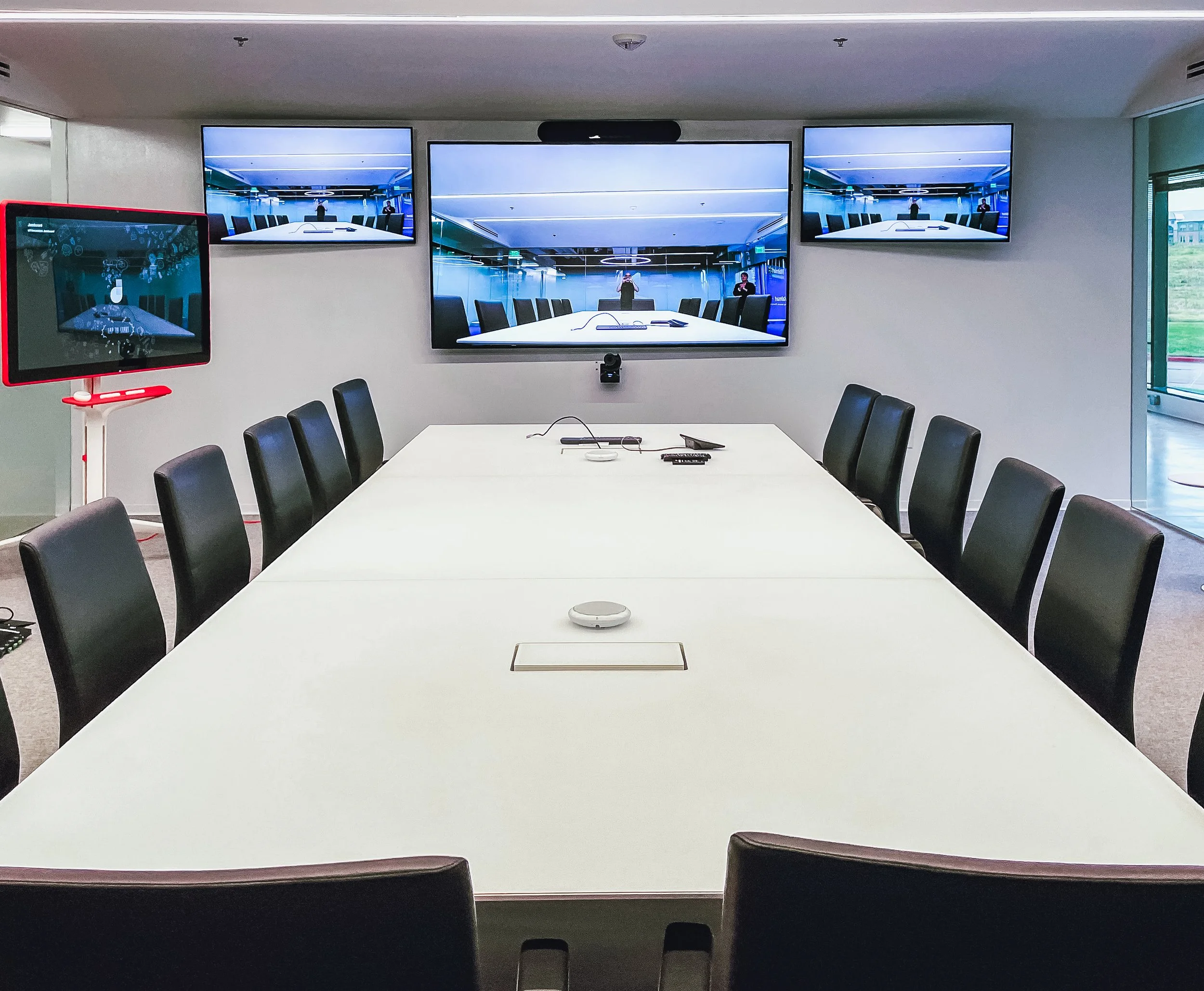 A conference room with a long white table surrounded by black chairs, three large screens on the wall displaying the same image of the conference room, a tablet on a stand to the left, and a window showing grass outside to the right.