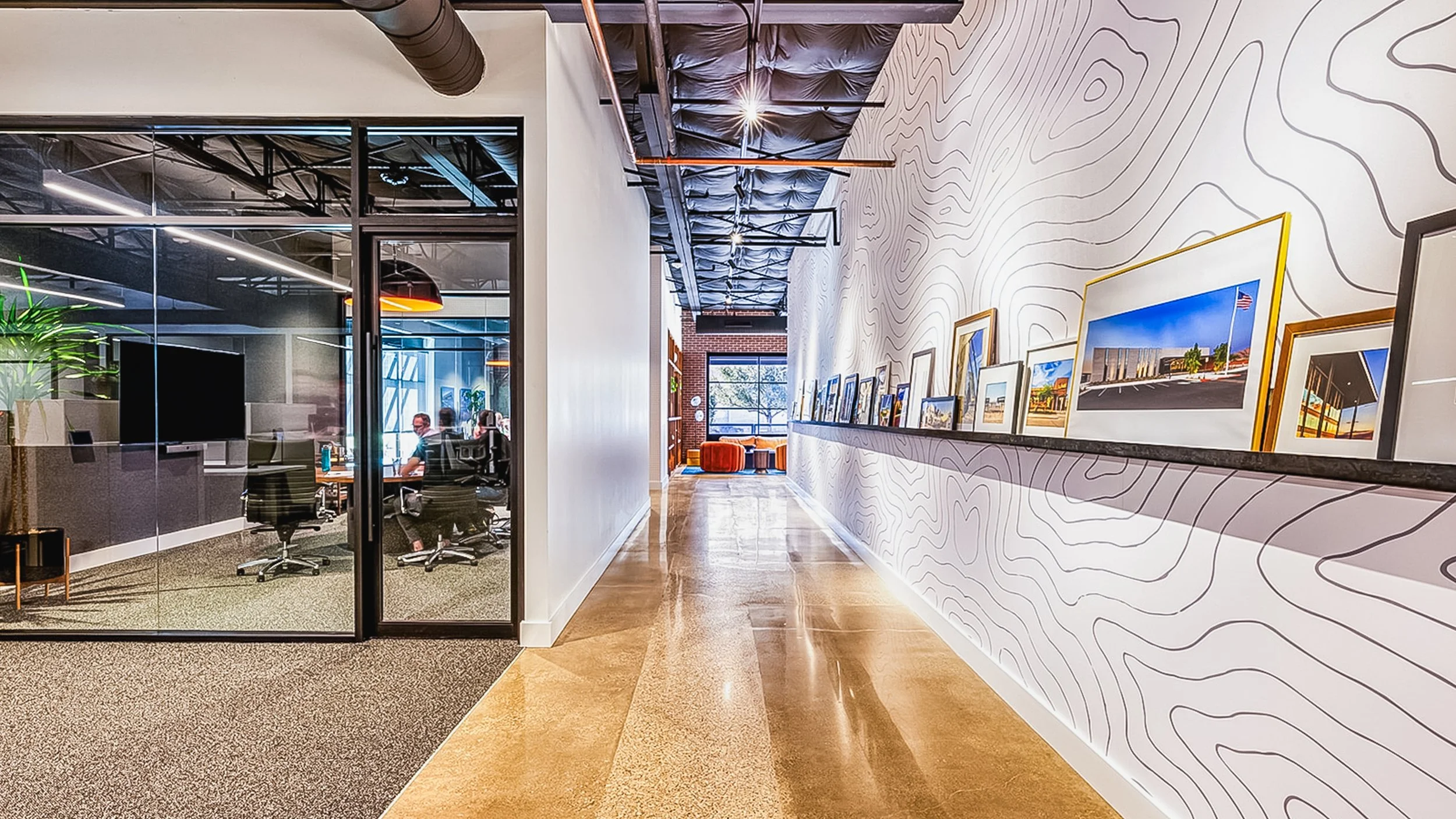 Modern office interior with a hallway, art display on the wall, and a glass-enclosed conference room with people working inside.