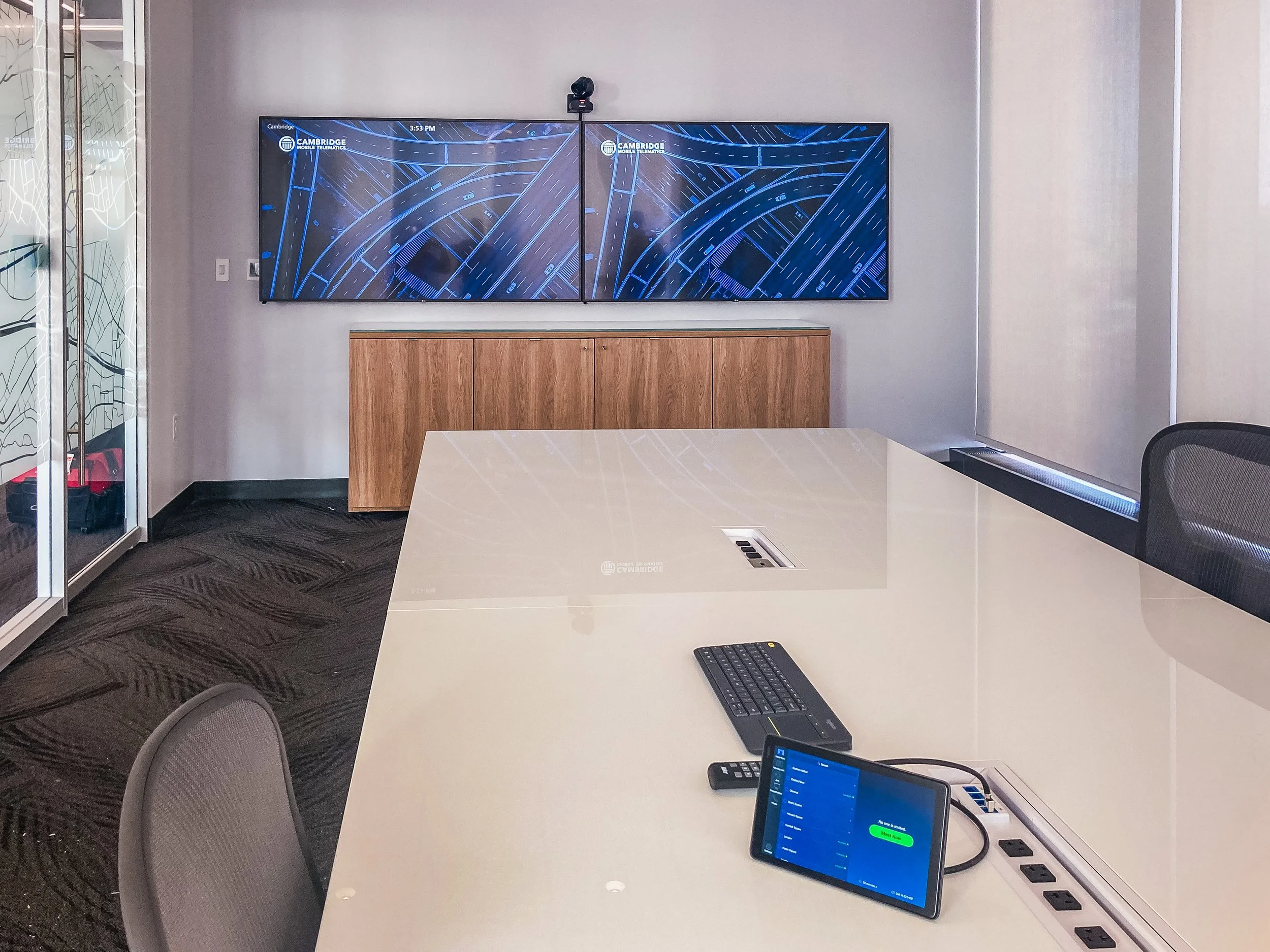 A conference room with a large white table, a wireless keyboard, a monitor, and a tablet displaying a screen with green text, a remote, and multiple power outlets on the side of the table. The background features two large digital screens showing a d