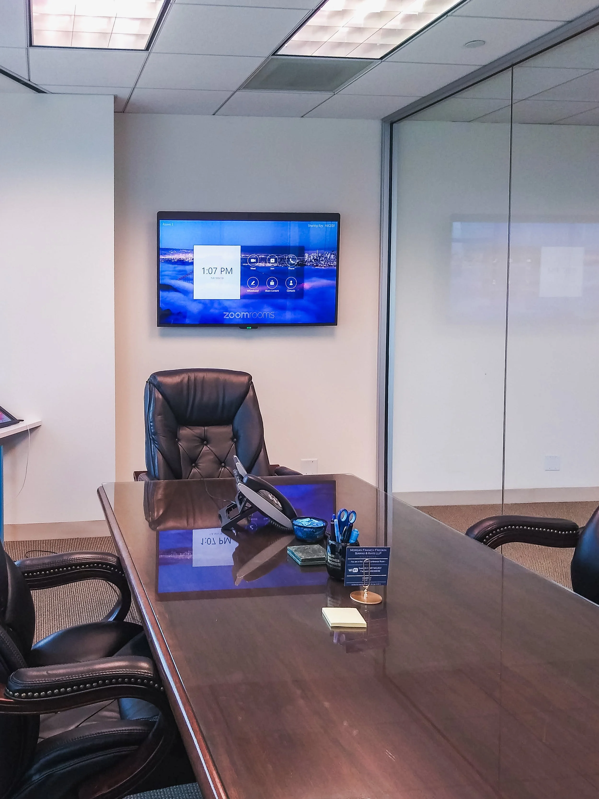 A conference room with a polished wooden table surrounded by black leather chairs. A large flat-screen monitor on the wall displays the time 1:07 PM and a cityscape background. The room is seen through a glass partition, with some office supplies and