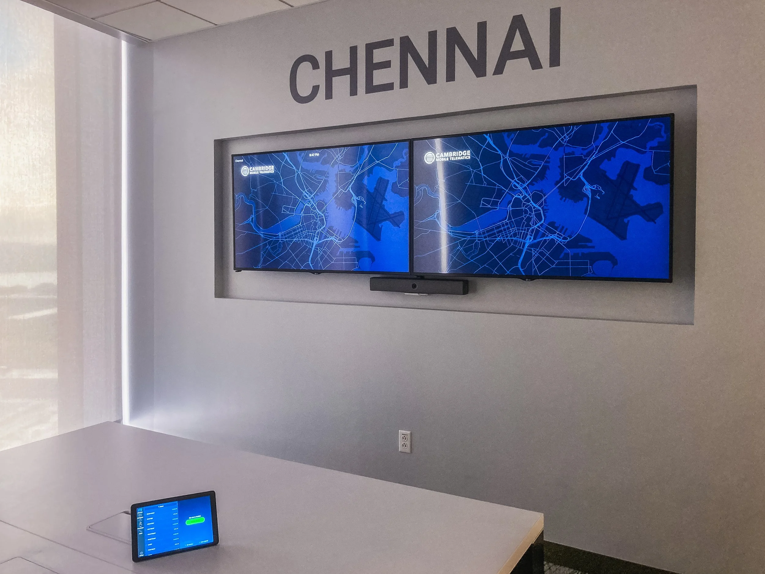 A room with a wall-mounted digital map display showing a city map of Chennai, with the word 'CHENNAI' above it. A small screen with a blue interface is on a desk below. The room has modern decor with a grey and white color scheme.