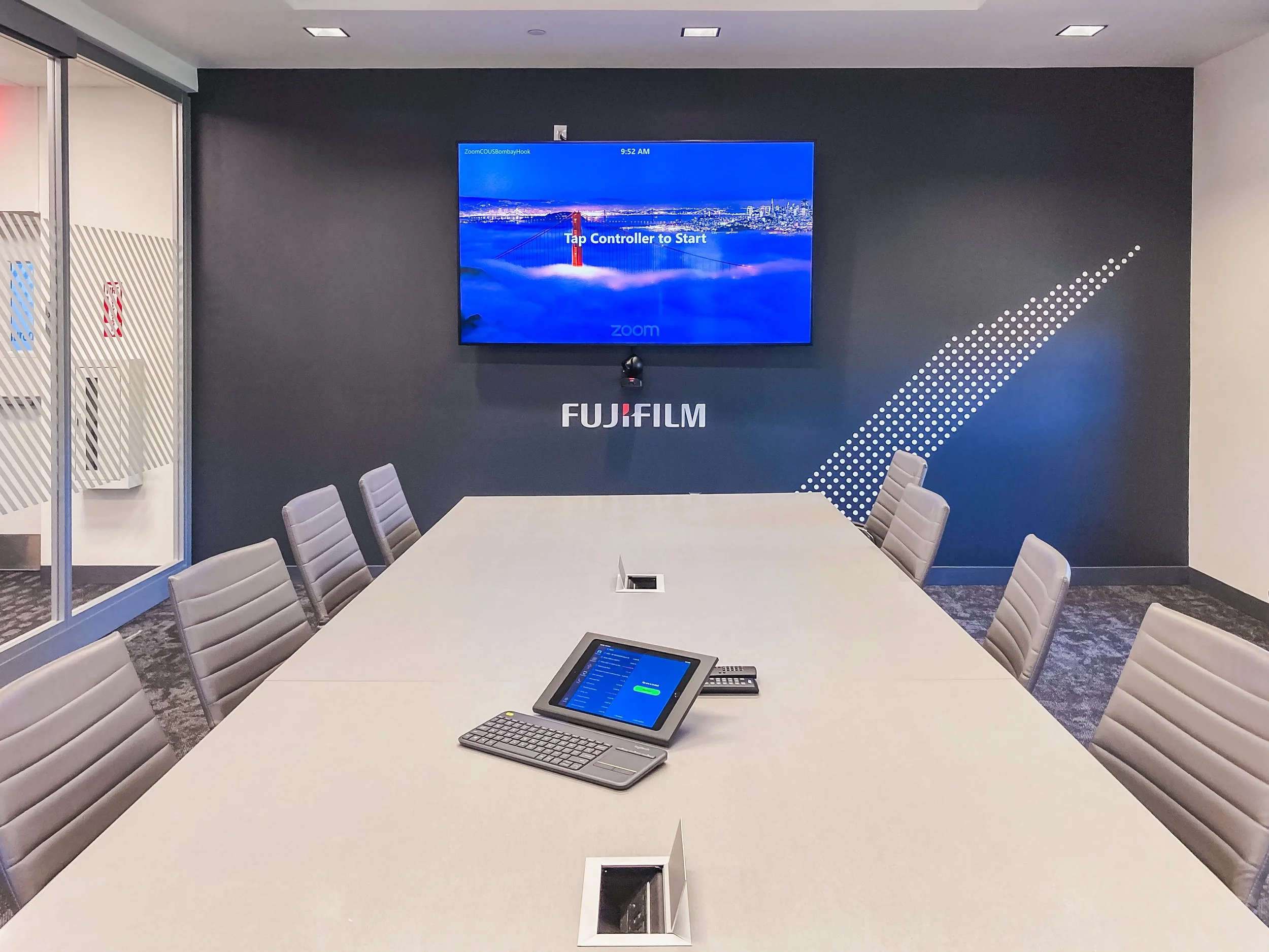 A conference room with a long table, chairs, a large wall-mounted screen displaying a cityscape and a prompt to tap a controller, and a tablet device with a keyboard and a telephone on the table.