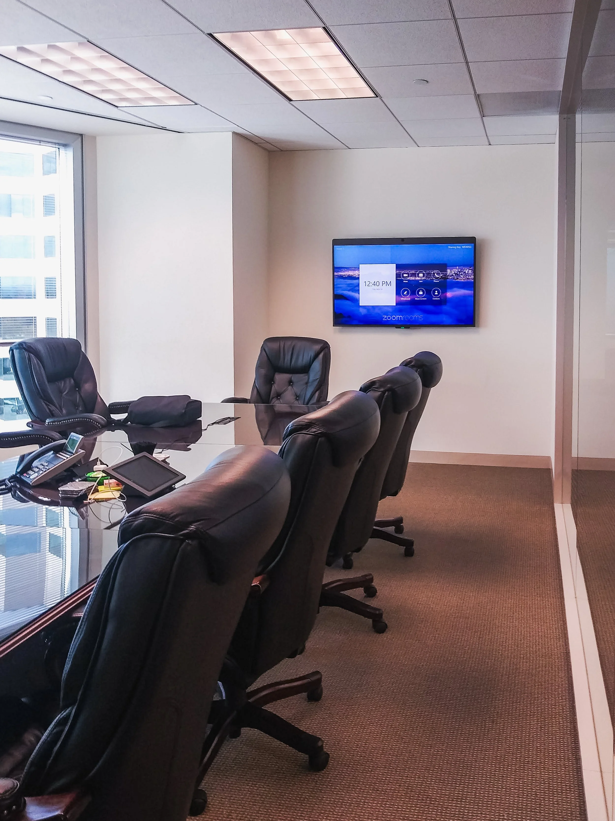 A conference room with a glass top table surrounded by black leather chairs, a wall-mounted monitor displaying a Zoom meeting interface, a window with city views, and a beige carpeted floor.
