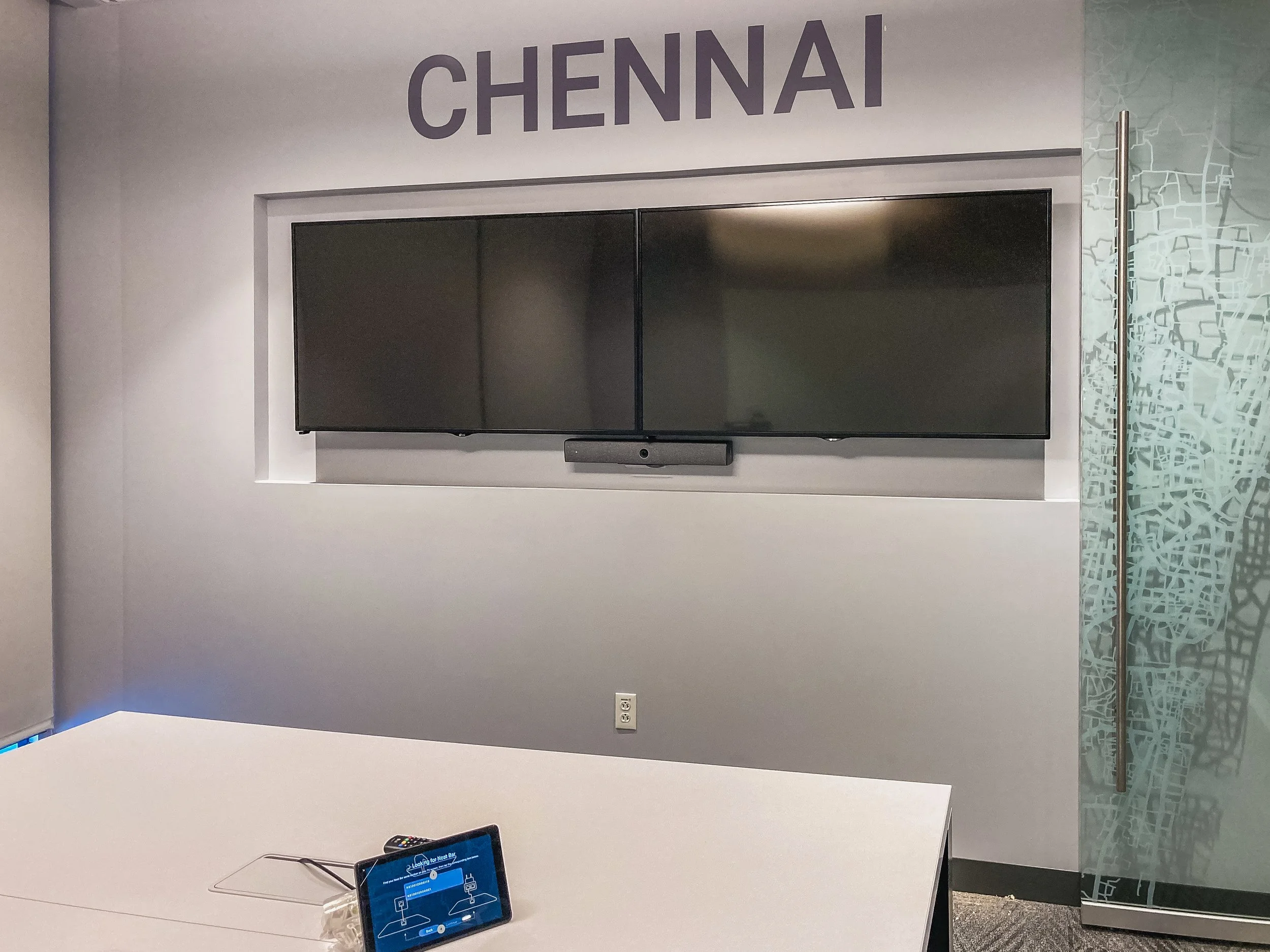 A large wall-mounted flat-screen TV in a room with the word 'CHENNAI' written above it, a soundbar below, and a small tablet device on a white table in the foreground. The room has white walls and a patterned glass partition to the right.