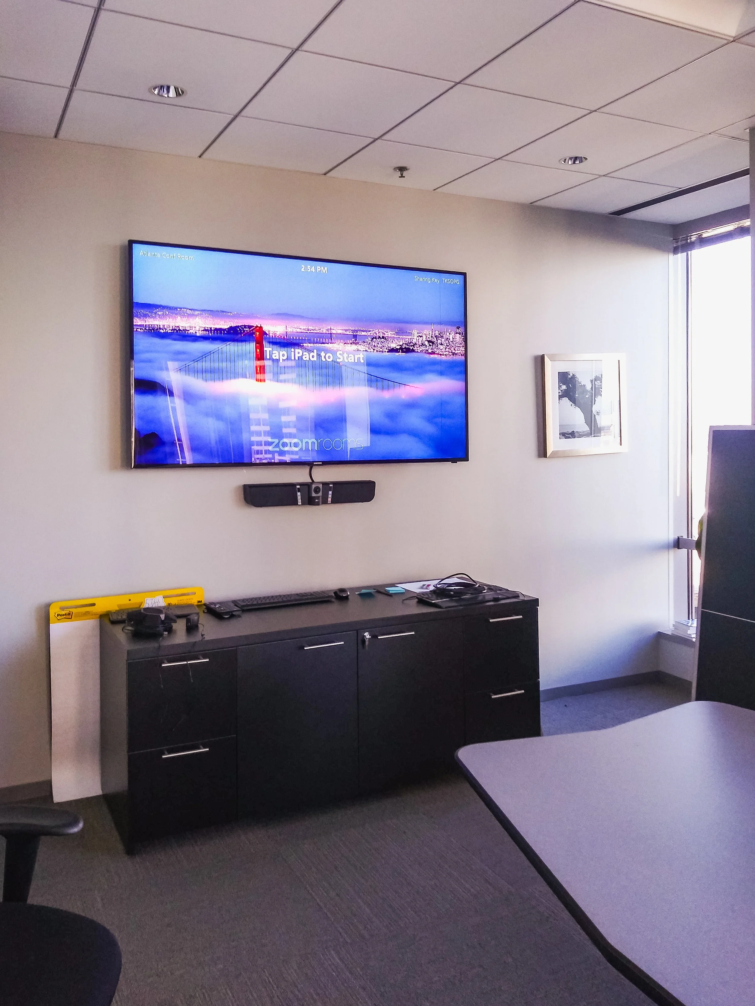 A conference room with a flat screen TV mounted on the wall displaying a city skyline and the words 'Tap iPad to Start'. There is a black cabinet below the TV with a remote, a yellow leveler, and some electronic devices on top. To the right, there is