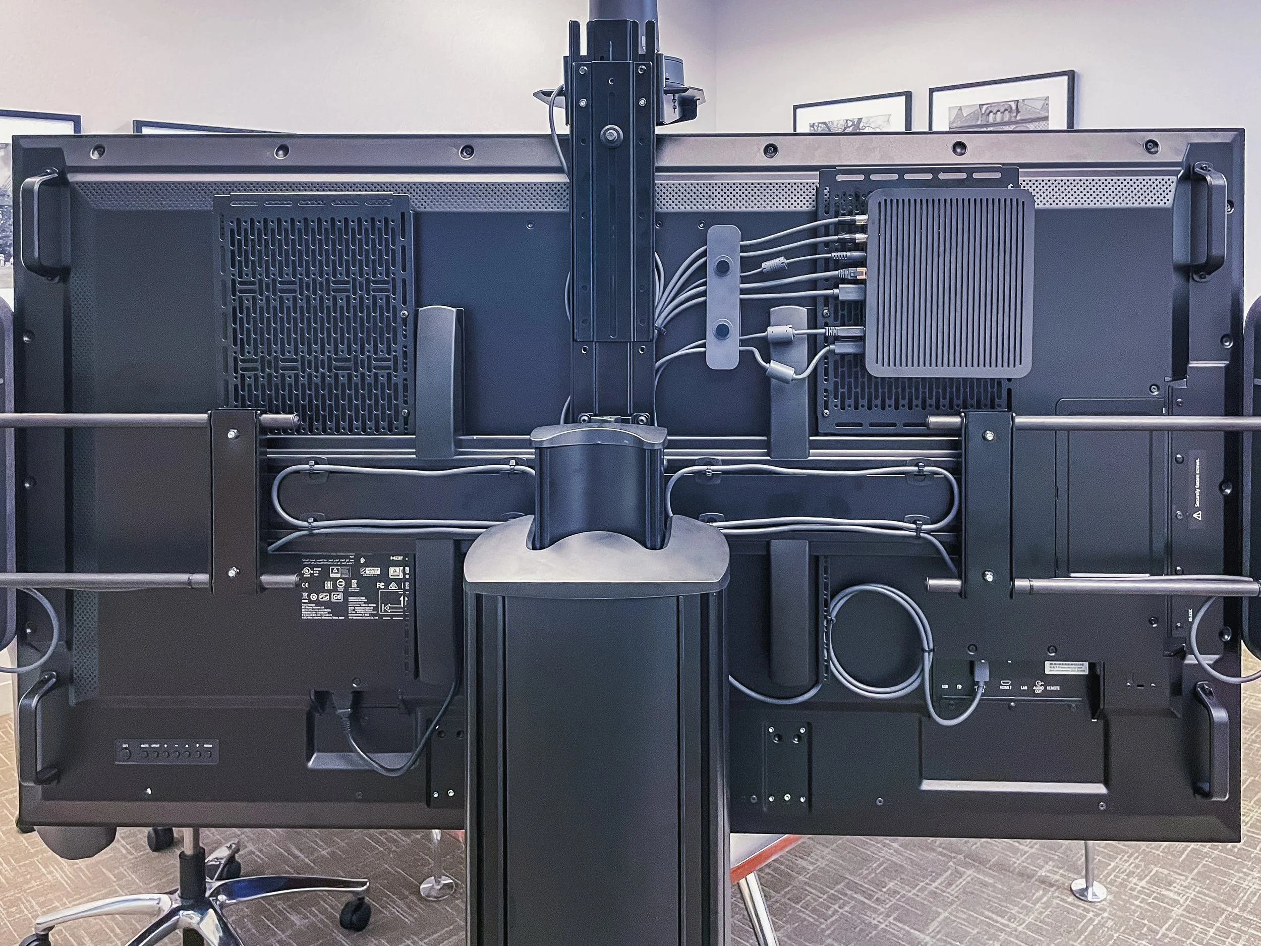 Rear view of a large flat-screen monitor on a black metal stand with multiple cables connected to a small gray device attached to the monitor.