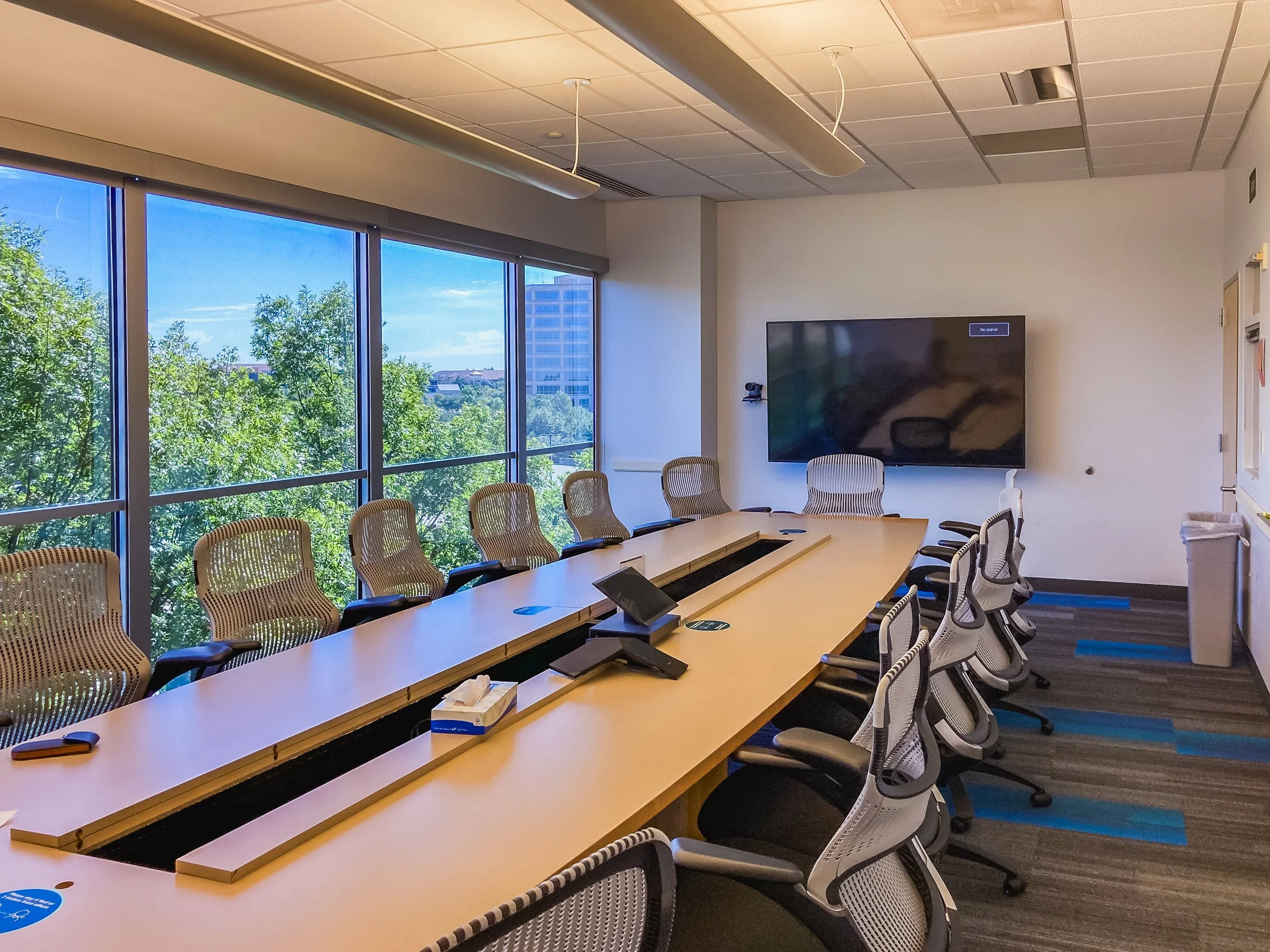 Conference room with a long wooden table surrounded by mesh office chairs. Large windows with a view of trees and a building outside. A large flat screen TV on the wall displays 'No signal.'