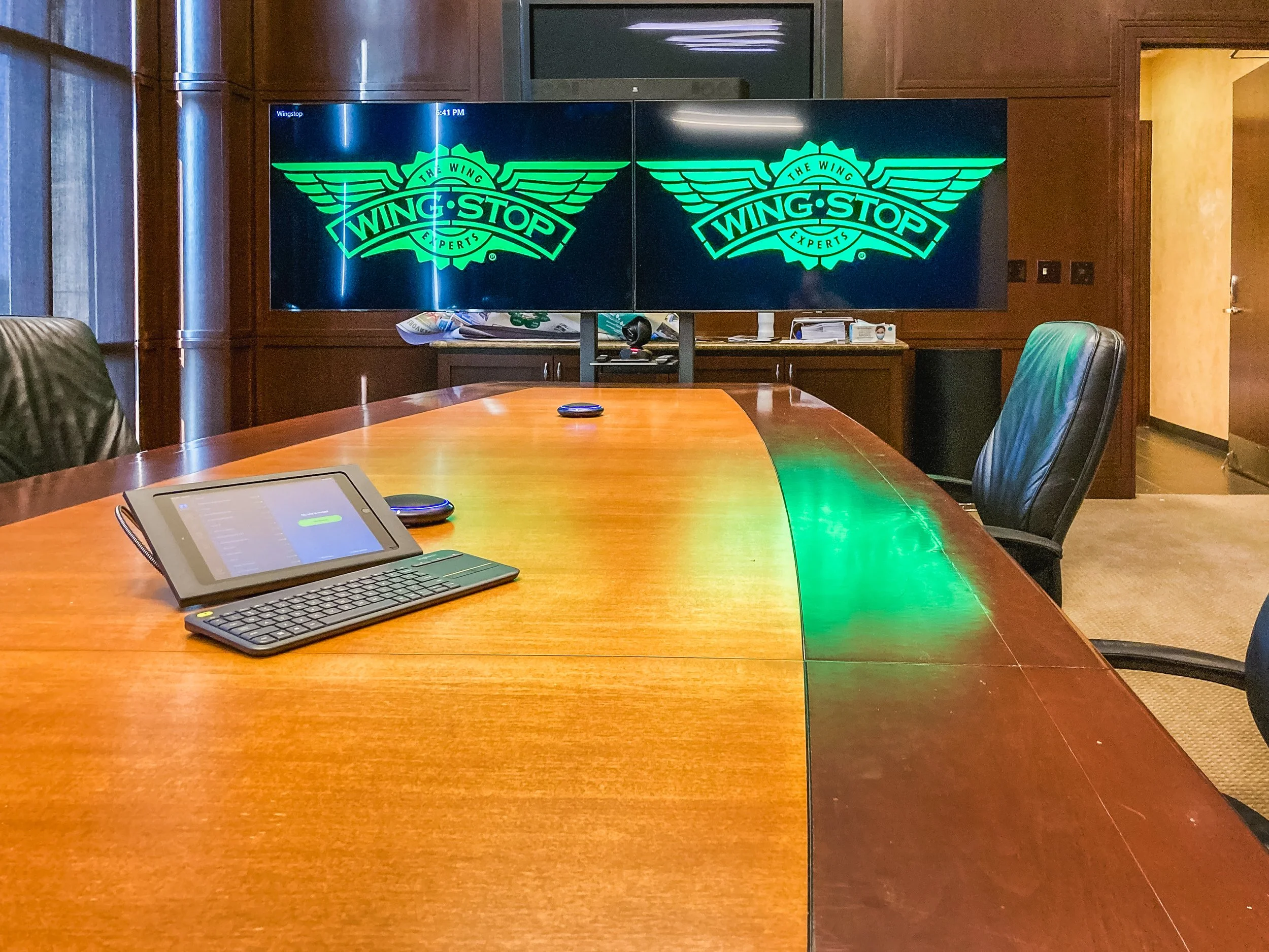 Conference room with a large wooden table, black chairs, and a wall-mounted screen displaying the Wingstop logo with green wings and text.