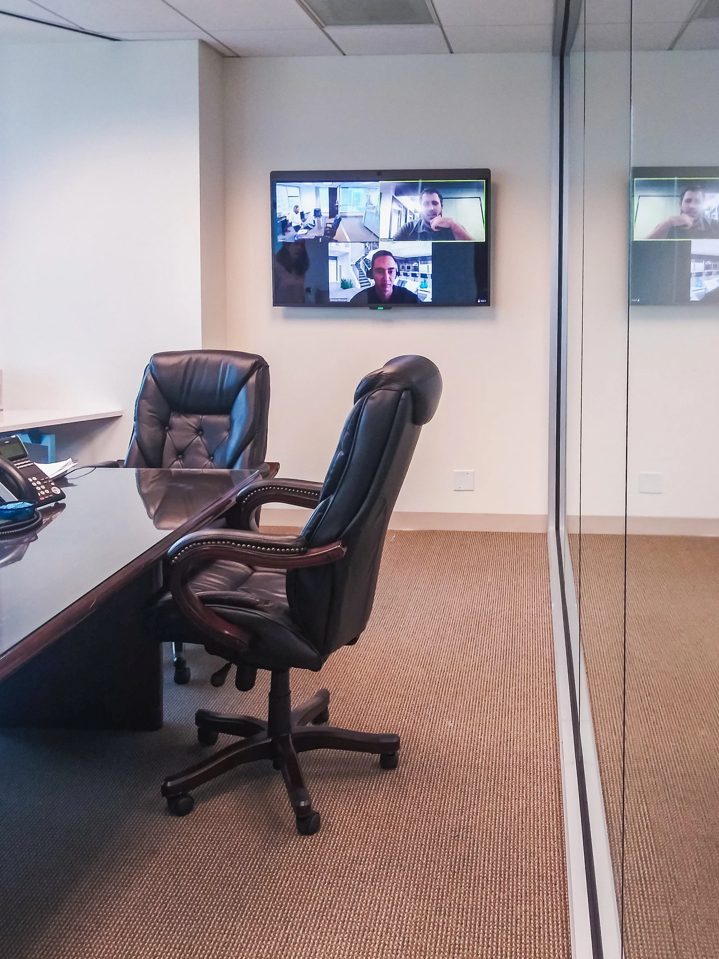 A conference room with a large wooden table, three black leather office chairs, a wall-mounted TV displaying a video call with three men, and a mirror on the right side reflecting part of the room.