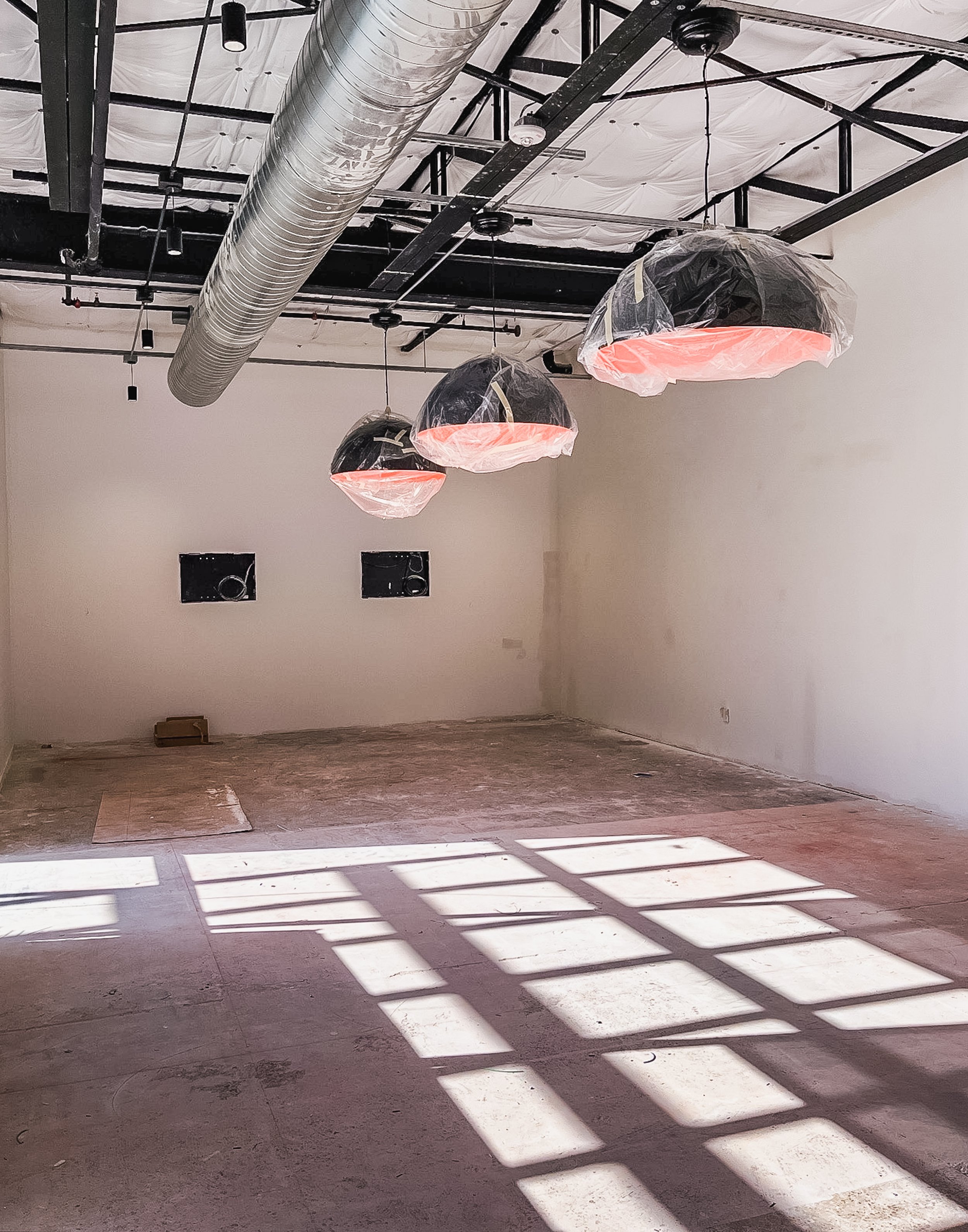 Empty room with unfinished flooring, hanging industrial-style lights with plastic coverings, large exposed ductwork on the ceiling, and shadows of window frames cast on the floor.