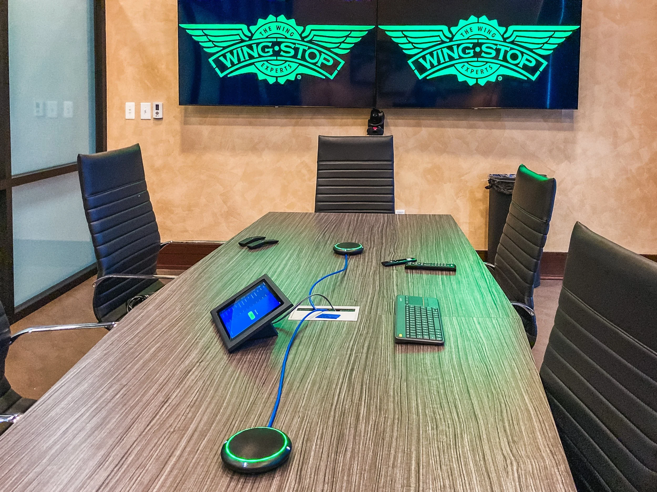 Conference room with a long wooden table, black leather chairs, a smart device, remote controls, and a glowing green smart speaker, with two large screens displaying the Wingstop logo on the wall.
