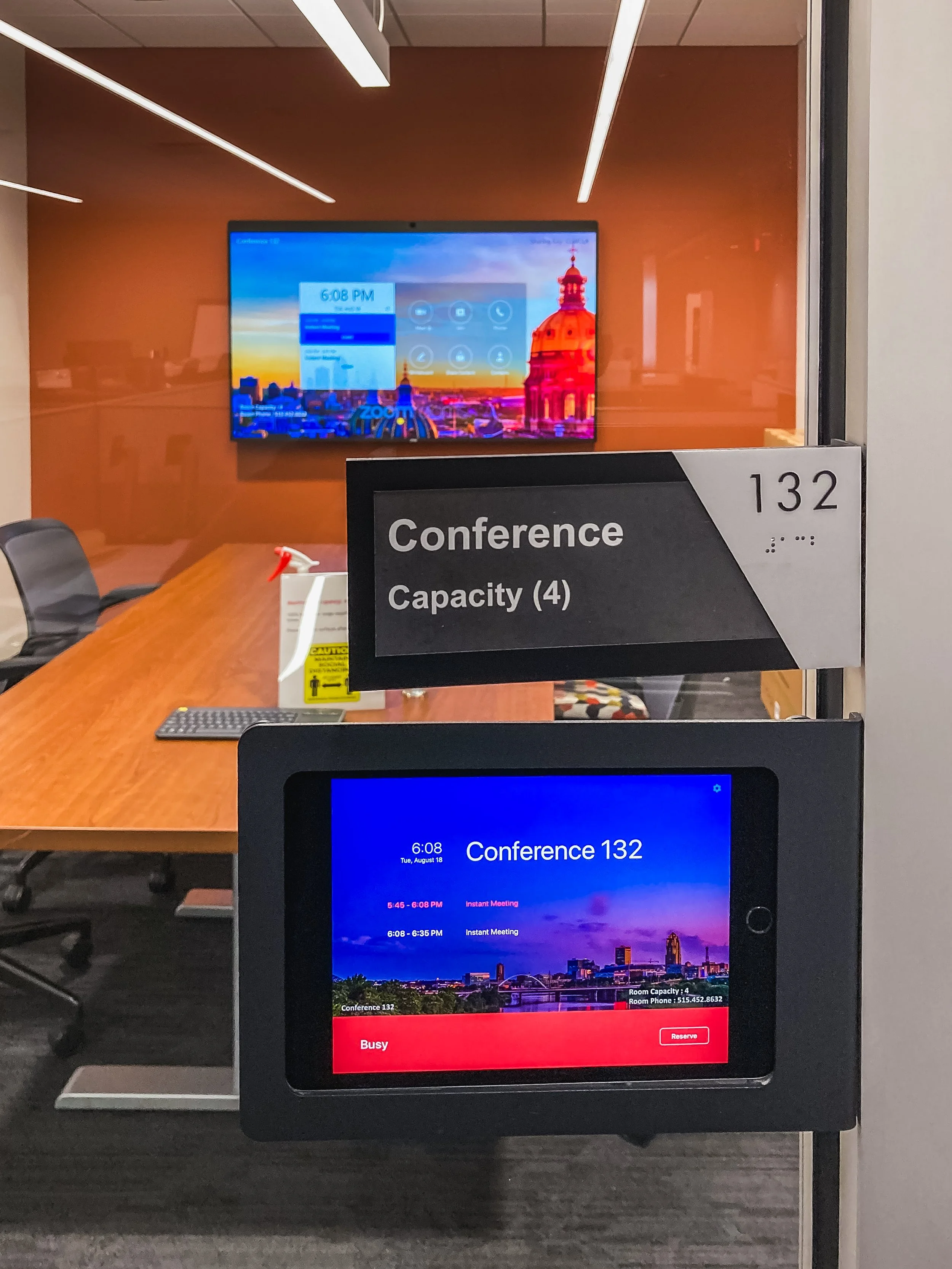 A conference room sign with the number 132 indicating room capacity of four, with a glass wall behind showing a conference room with chairs, a table, a monitor, a spray bottle, and a chair, and digital screens displaying meeting schedules.