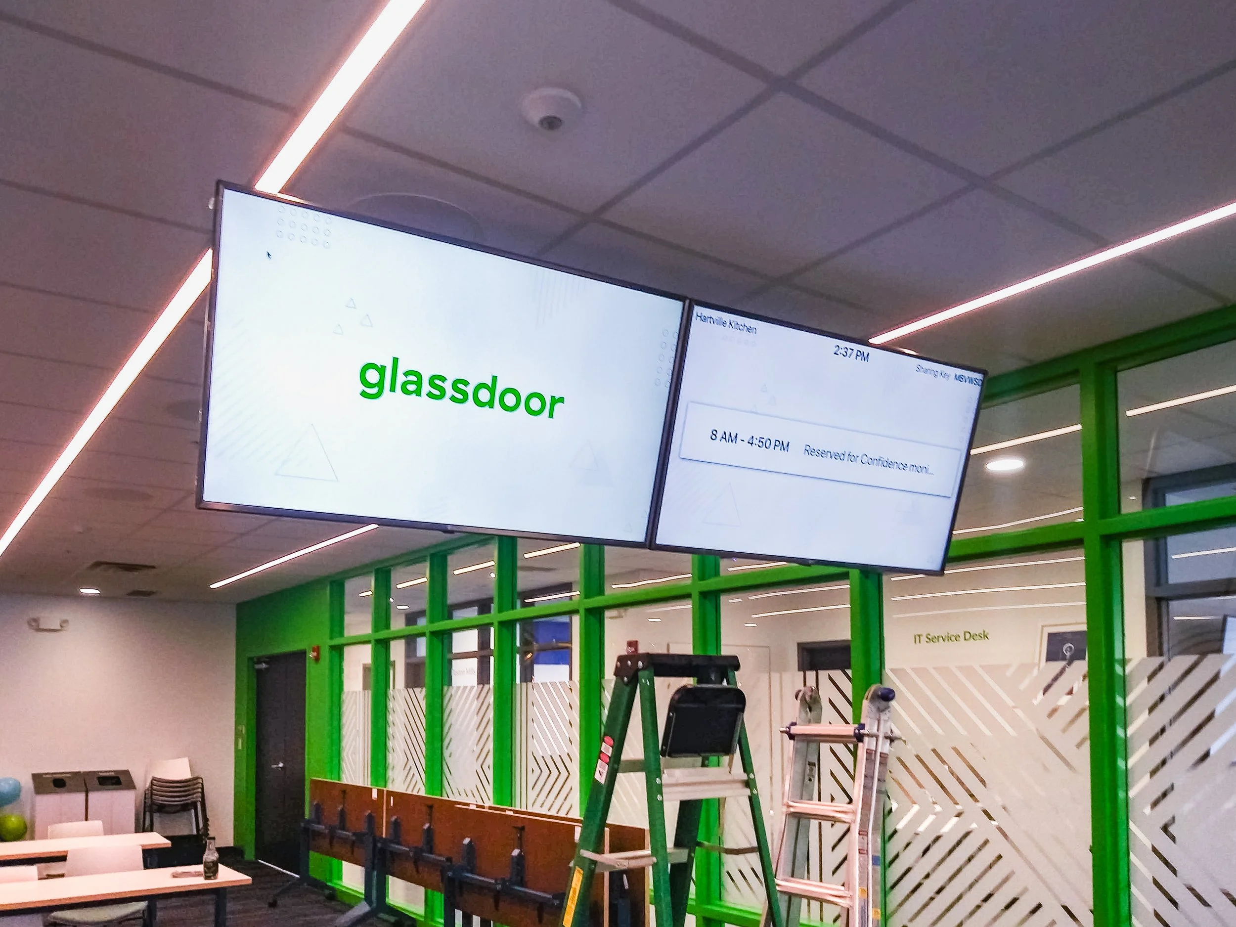 Large digital screens displaying text, one says 'glassdoor' and another shows reservation details, in a modern office or waiting area with green structural elements, furniture, and equipment including a ladder and exercise bike.