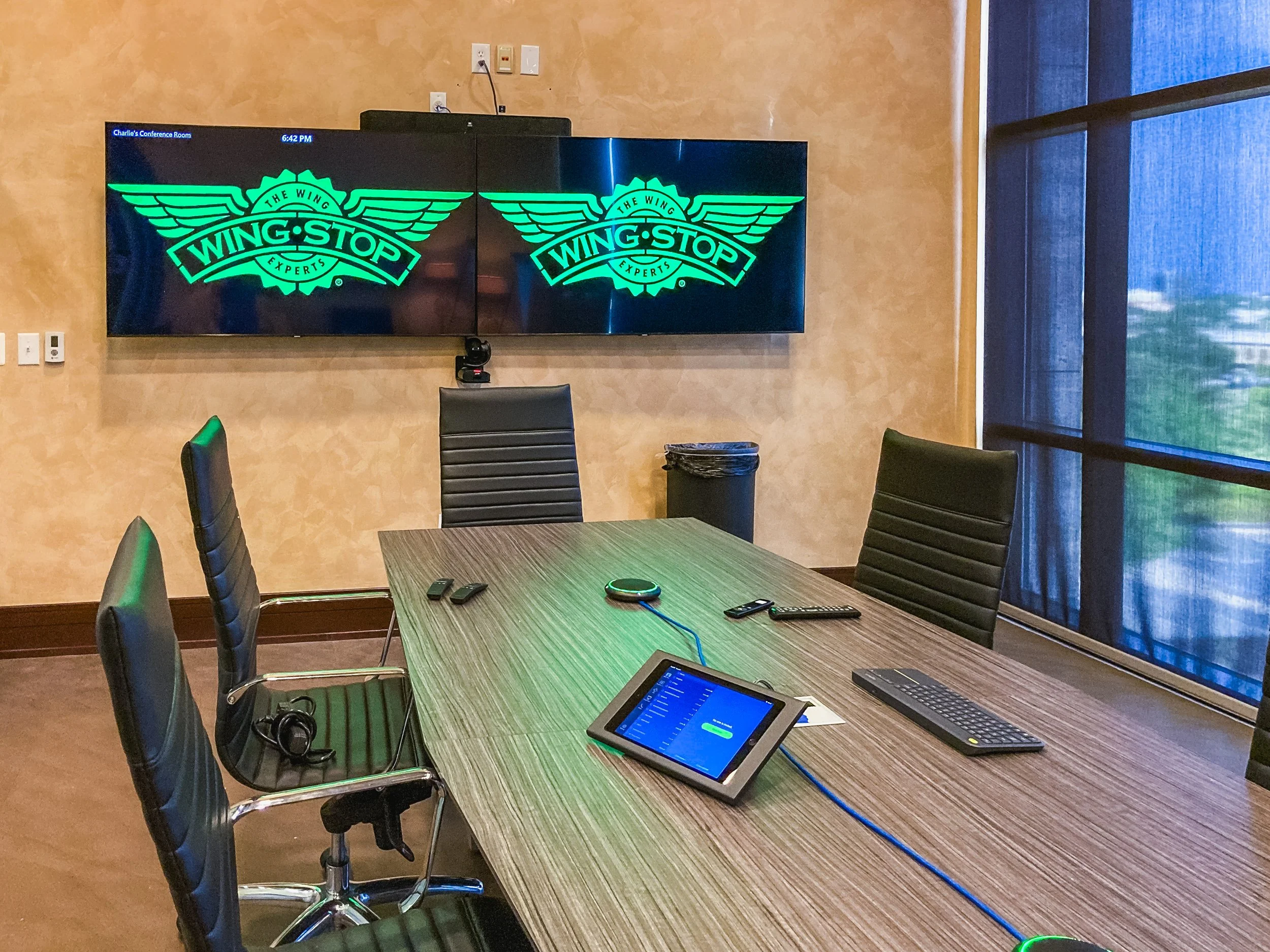 A conference room with a large wooden table, black chairs, a wall-mounted TV displaying a logo for Wing Stop, and a tablet, keyboard, and remote control on the table. The room has a window with blue curtains and yellow textured walls.