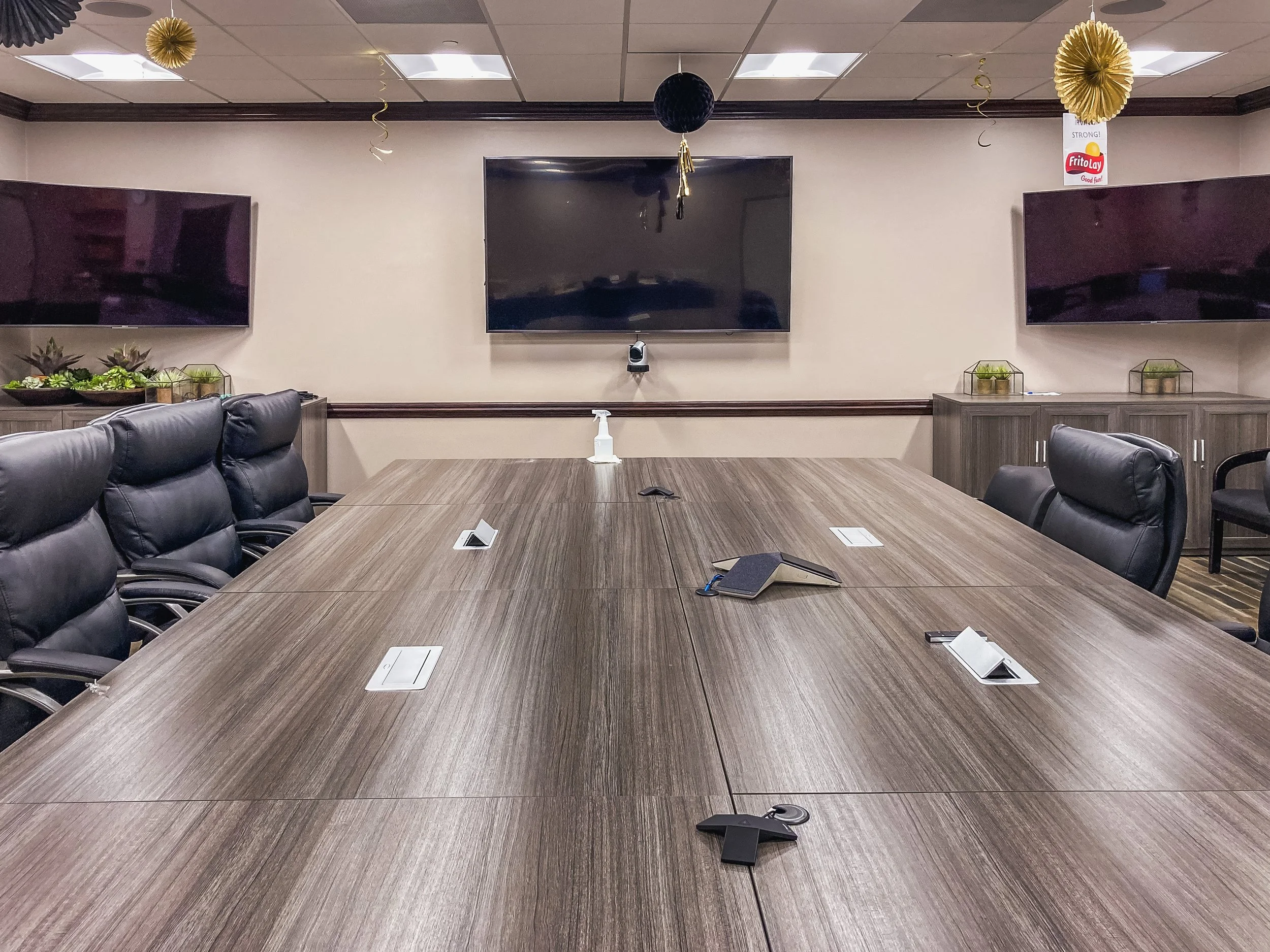 A conference room with a long wooden table surrounded by black leather chairs. There are two large TV screens on the wall, a zoom camera beneath the middle screen, and a bottle of hand sanitizer on the table. Decor includes gold and black hanging orn