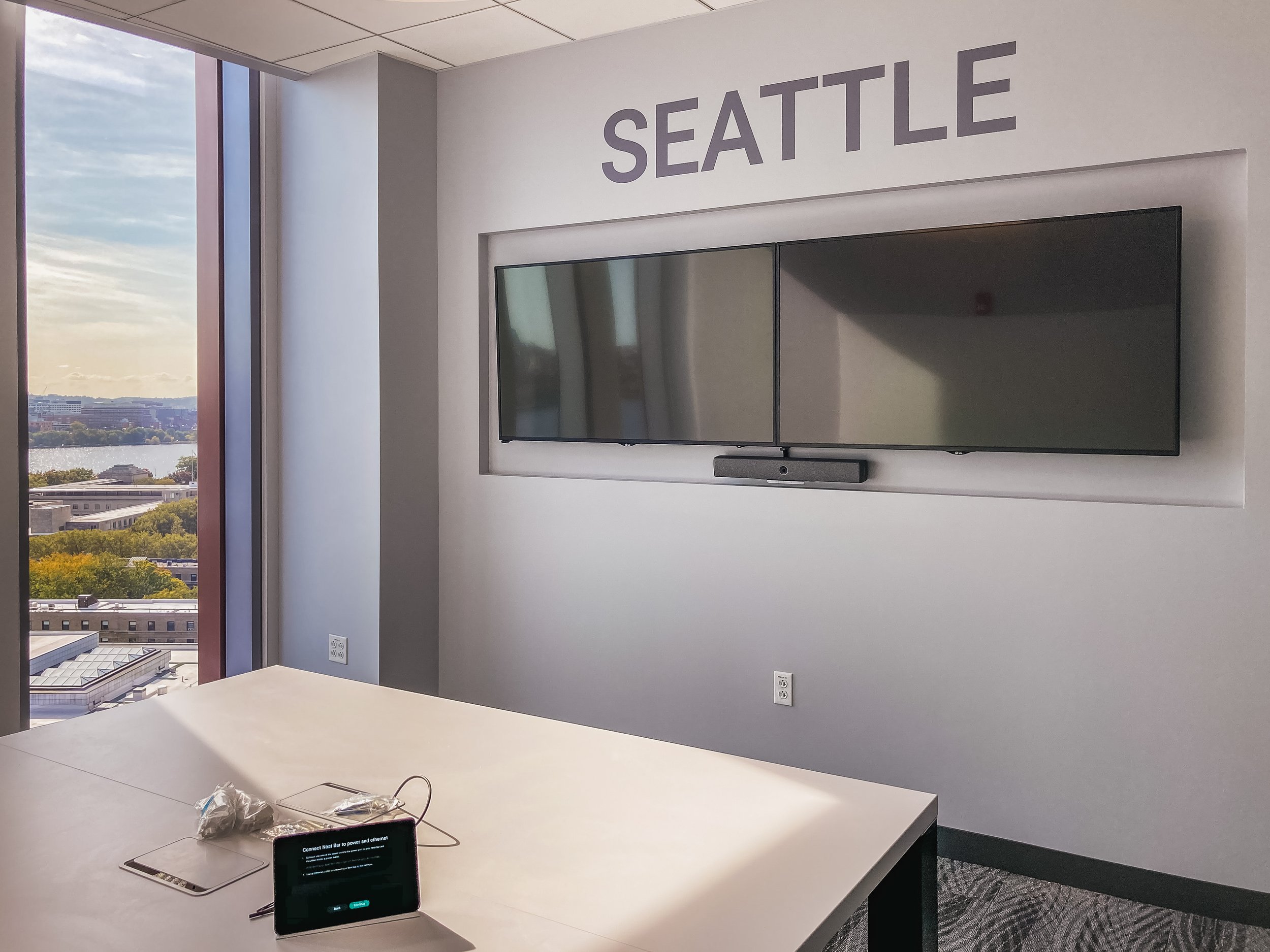 Office conference room with a large window showcasing a city and water view, a white table with electronic devices and cables, and a wall-mounted TV screen near the Seattle sign.